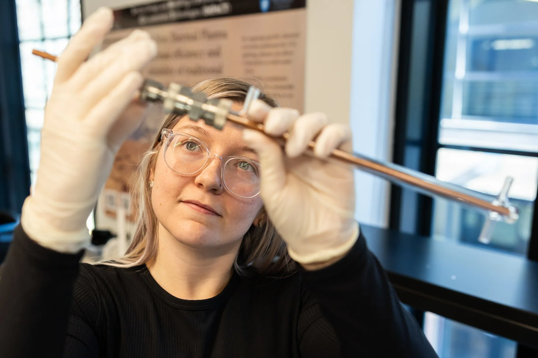 A woman wearing glasses and latex gloves handling a copper tube with a valve in a laboratory or workshop setting.