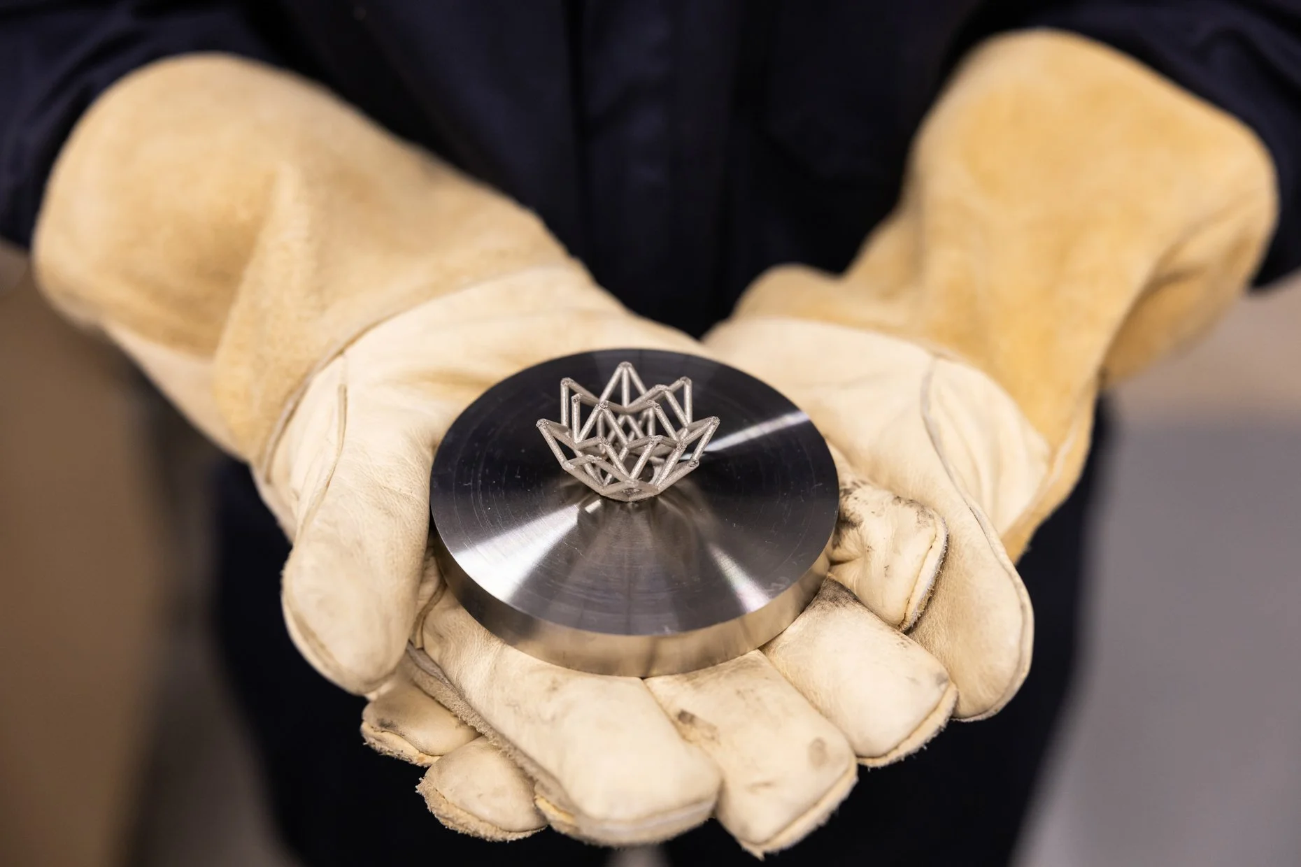 A person wearing beige work gloves holding a circular metal disc with a small geometric metallic sculpture on top.