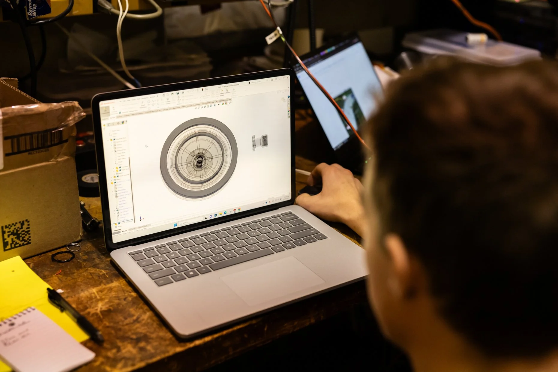 A person working on a laptop with a detailed technical design or technical drawing displayed on the screen. The workspace includes a cardboard box, a notepad, a yellow note, and some scattered small items on a wooden table.