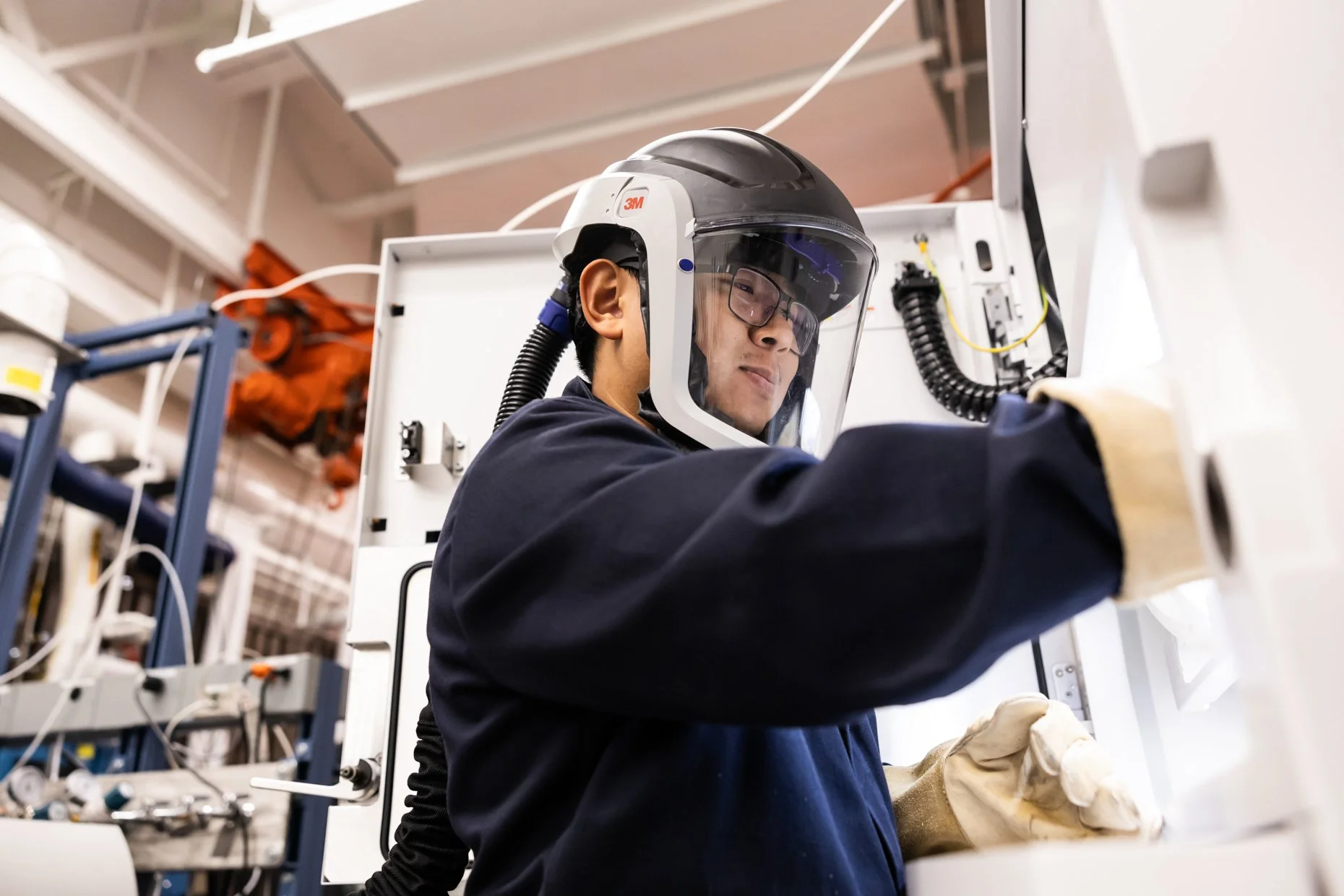 A worker wearing a 3M helmet, safety goggles, and gloves working with machinery in an industrial setting.