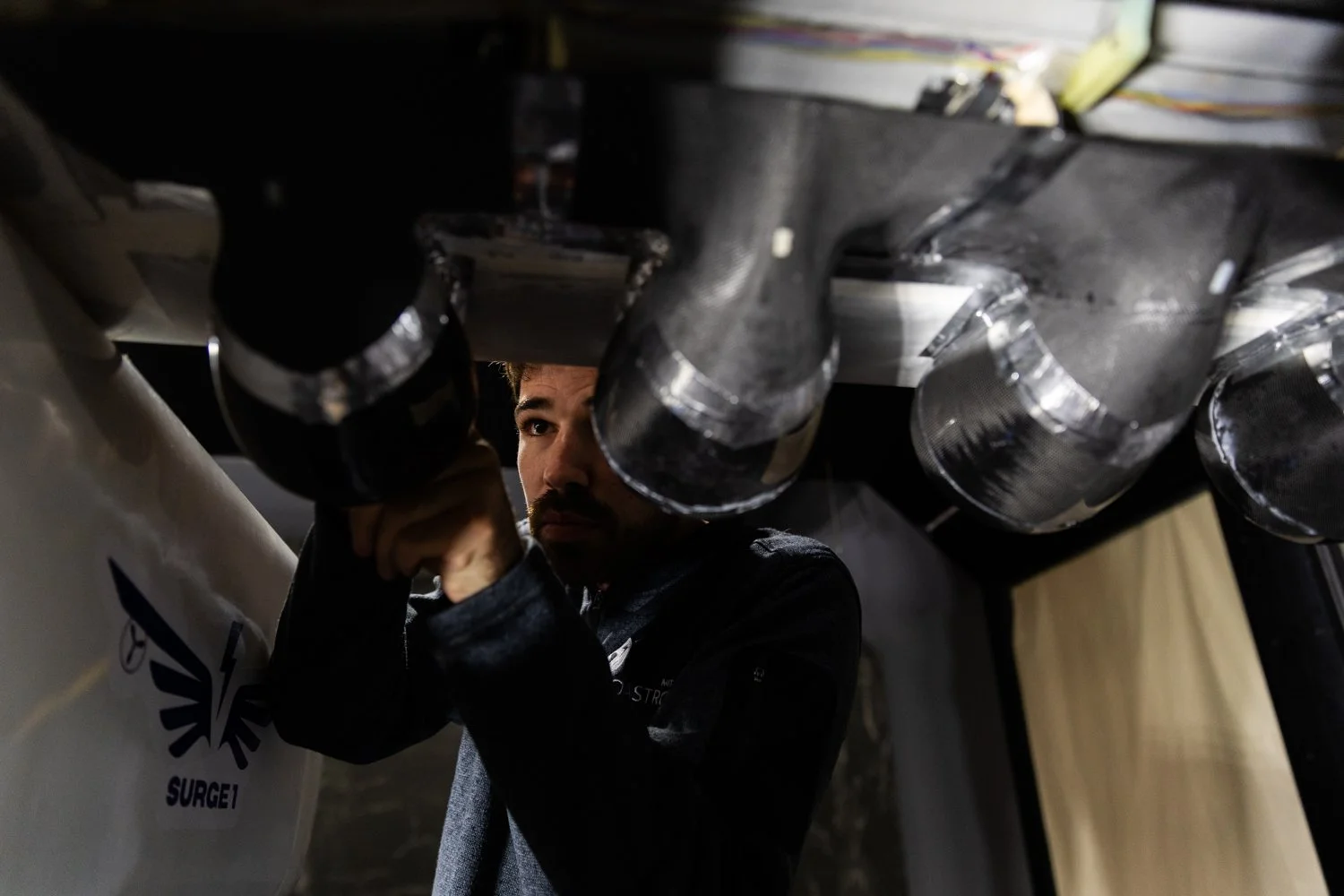 A man working on a mechanical or electrical component beneath a structure with several cylindrical pipes wrapped in reflective insulation material.