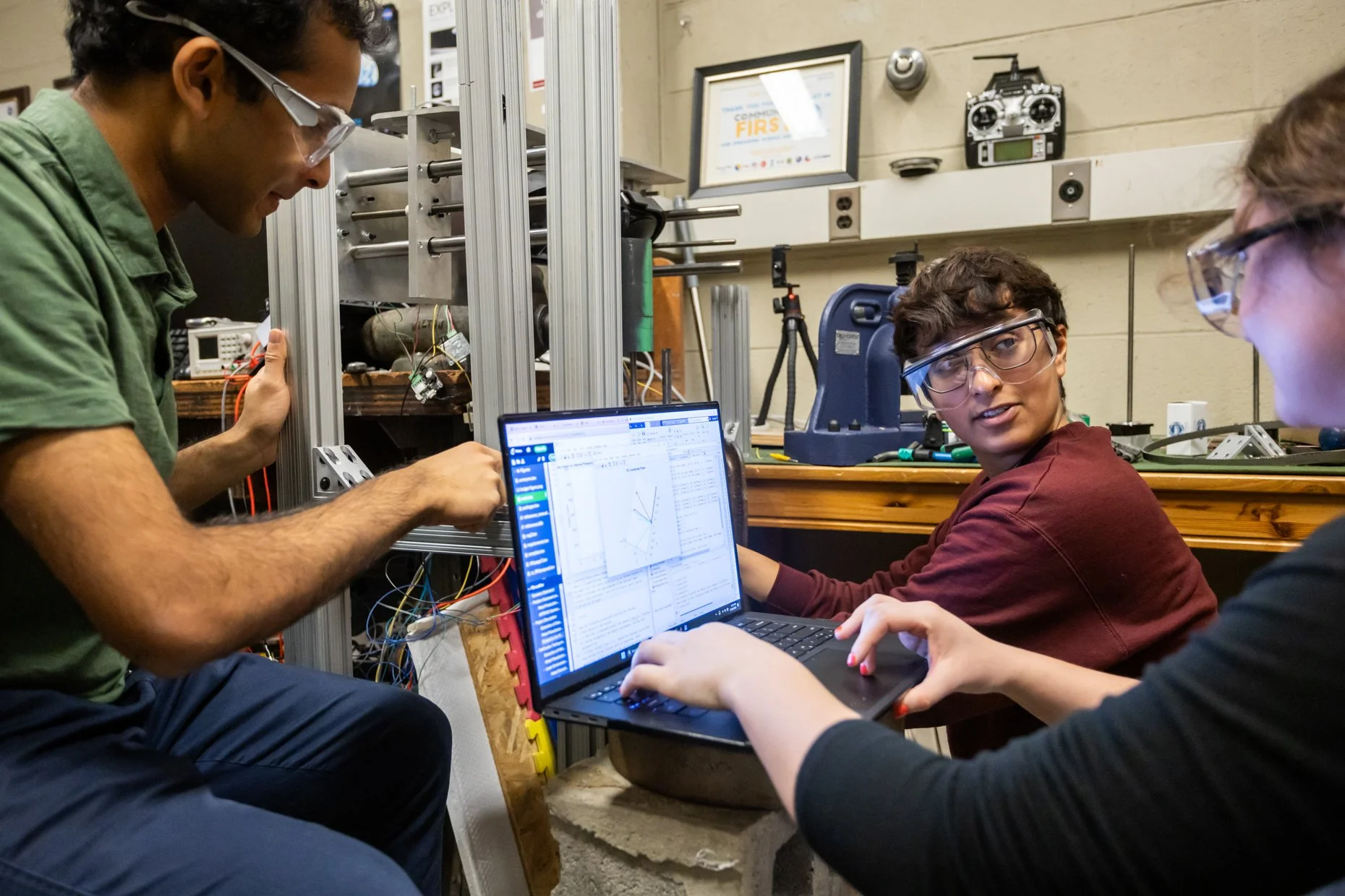 Group of three students working on robotics project in a workshop, with one using a laptop displaying a graph and two others adjusting the robot with wires and tools.