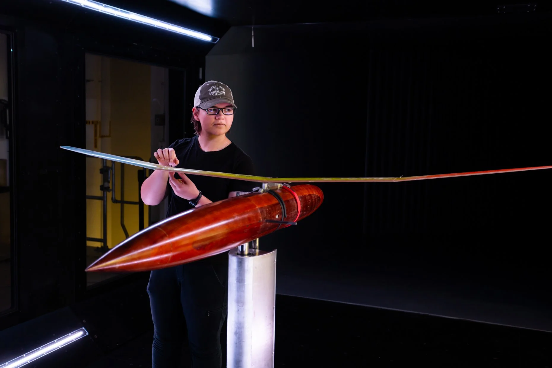 A person wearing glasses and a gray cap is adjusting a large, spinning object that resembles a torpedo or missile, mounted on a stand indoors with black walls and minimal lighting.