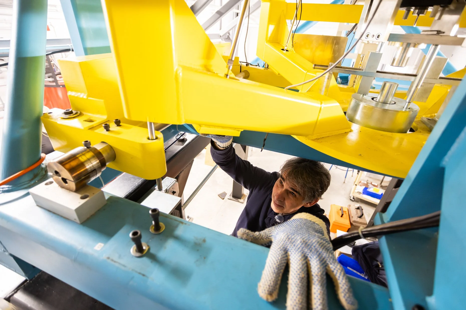 A man working on a large blue and yellow mechanical structure, using tools and wearing protective gloves. The structure appears to be part of industrial machinery or equipment.