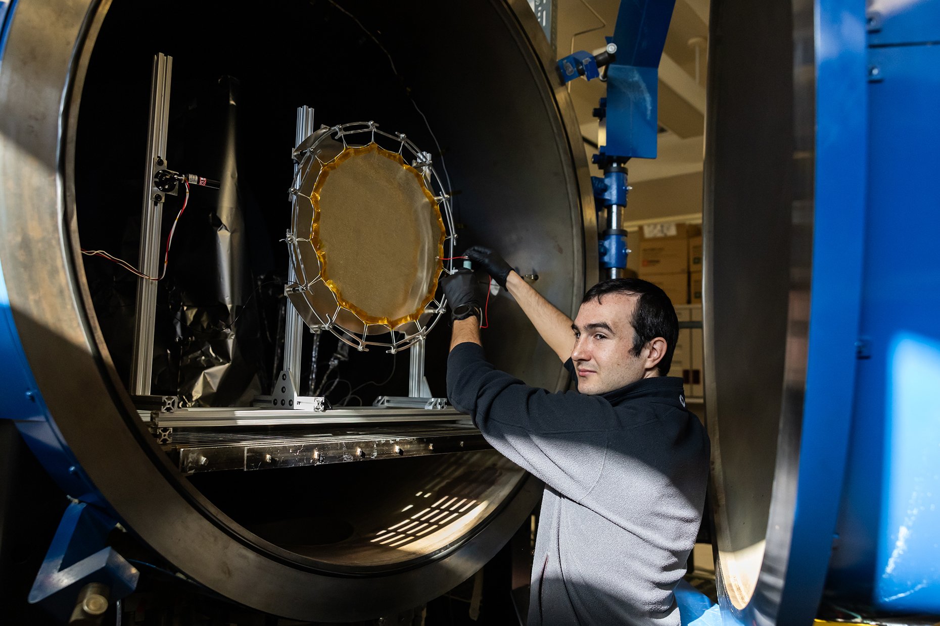 A scientist working inside a large vacuum chamber with a circular object, likely a scientific instrument or component, mounted on a metal frame. The scientist is adjusting the setup and wearing gloves, with various equipment and structural elements v
