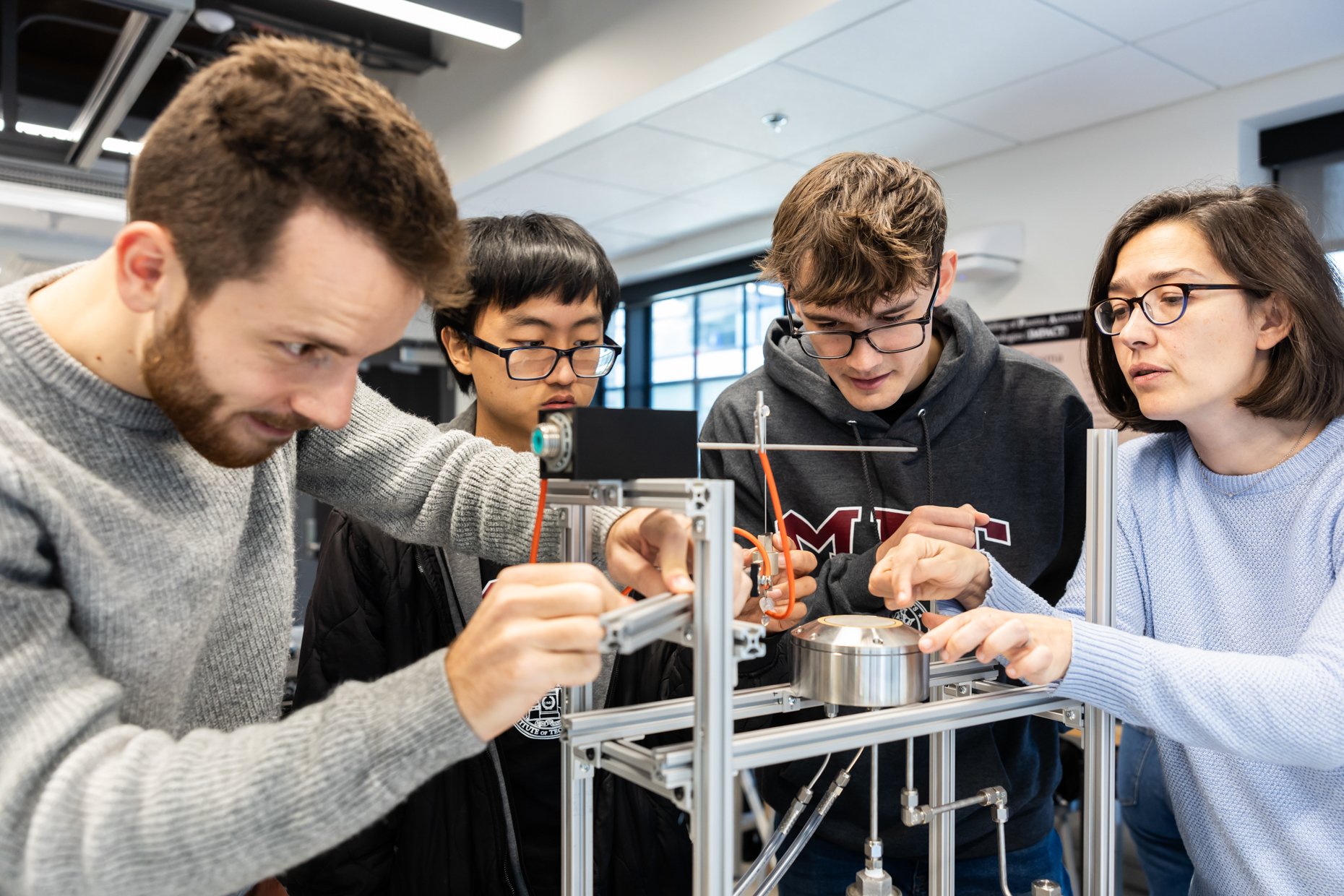 Group of four students working on a scientific experiment with equipment and machinery in a laboratory or classroom setting.