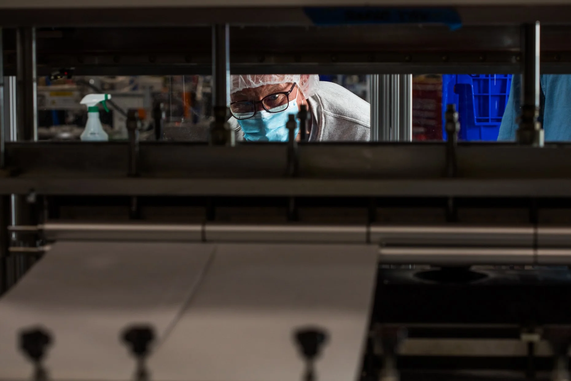 A person wearing glasses, a face mask, and a hairnet looking through a metal rack in a factory or laboratory setting.