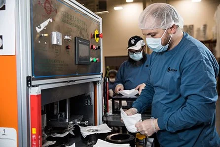 Two workers in hairnet, mask, and blue uniforms working with a machine in a laboratory or manufacturing setting.
