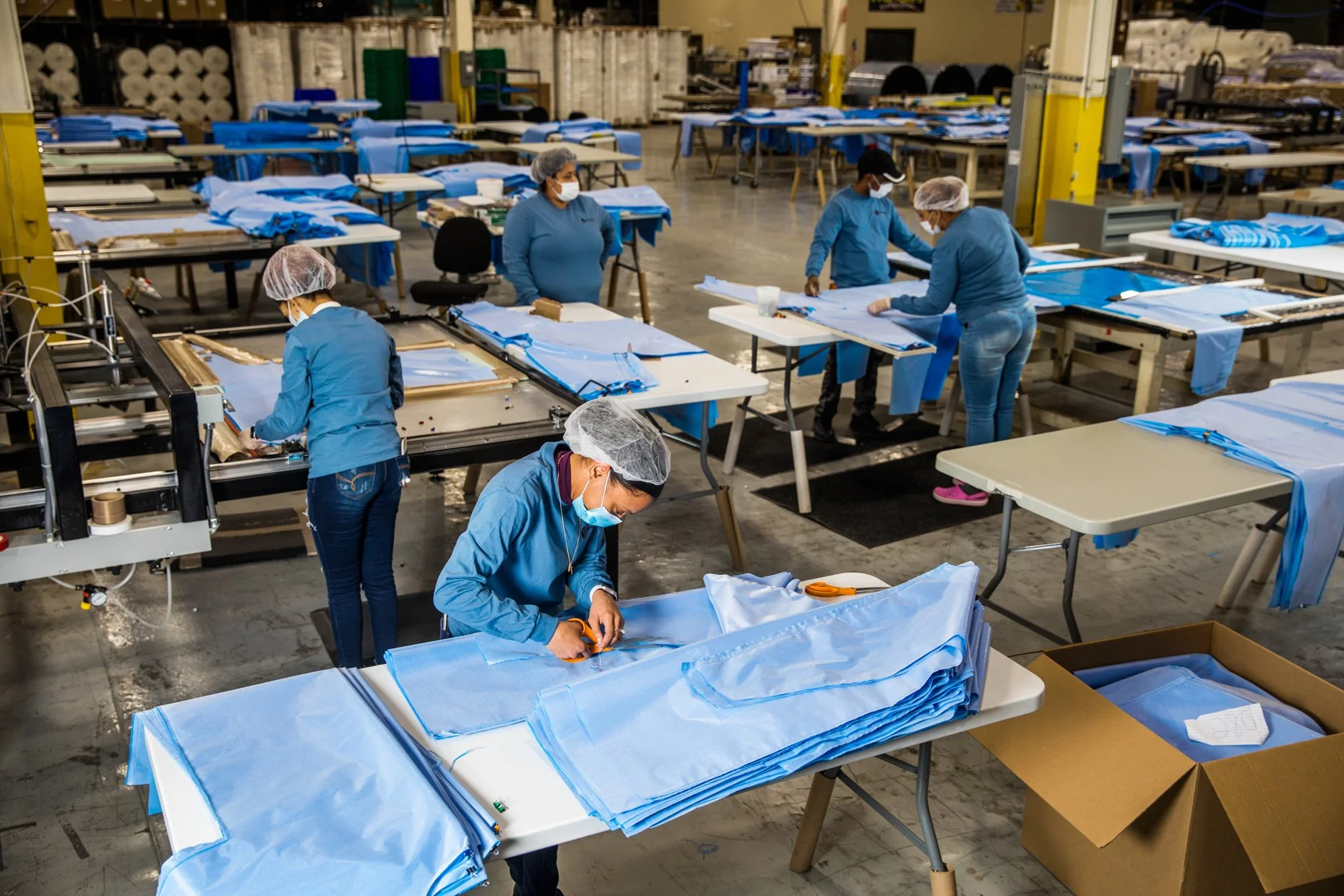 Workers in a factory sewing blue fabric, wearing masks, hairnets, and gloves, with tables, fabric, scissors, and sewing machines in a large industrial workspace.