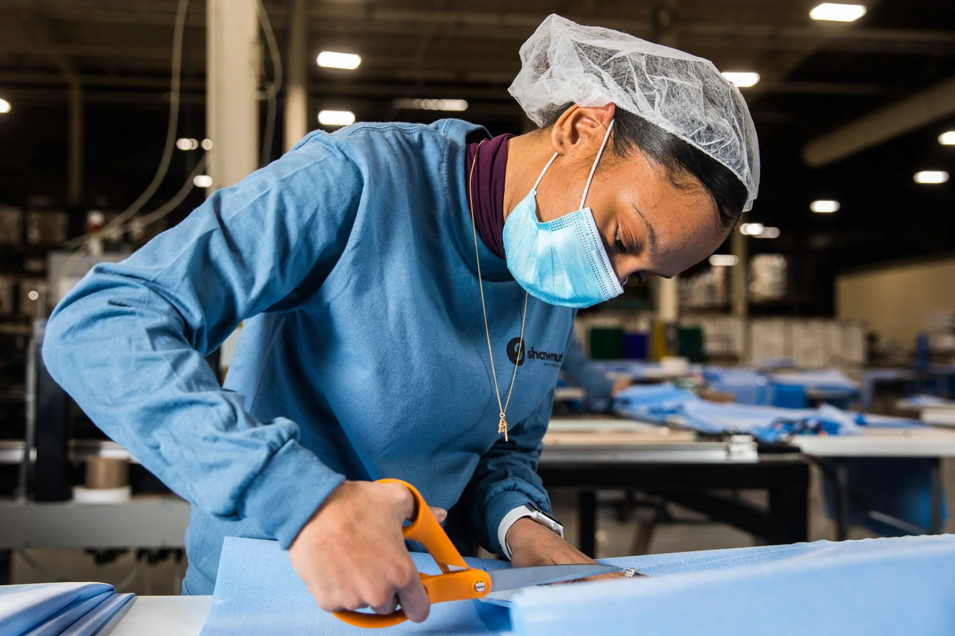 A woman wearing a blue shirt, surgical mask, and hair net cuts a piece of blue fabric with orange scissors in a factory or workshop setting.