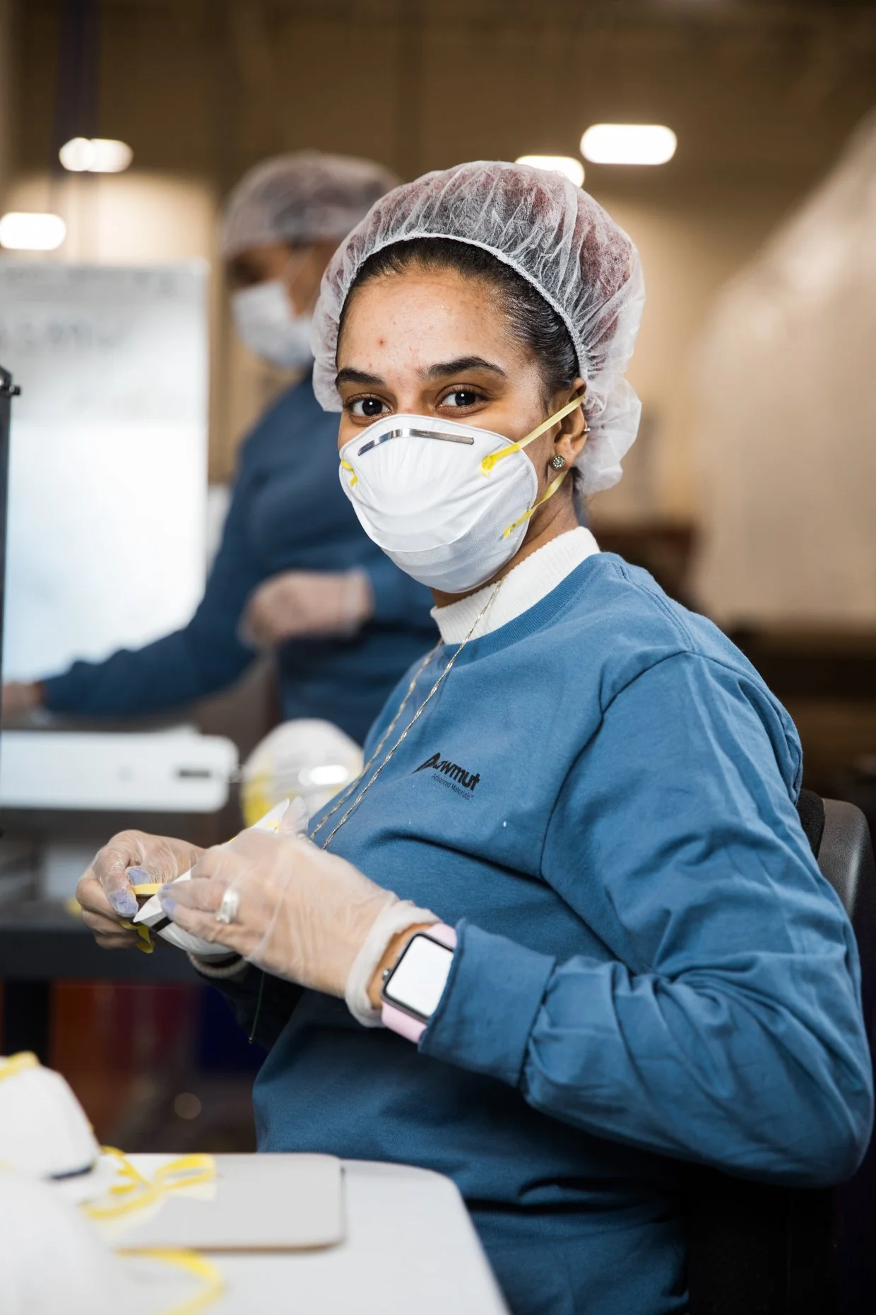 A woman wearing a blue medical scrubs, a surgical cap, a N95 mask, and gloves, working on a medical device or equipment in a clinical or manufacturing setting. She is smiling and looking at the camera, with another person blurred in the background.