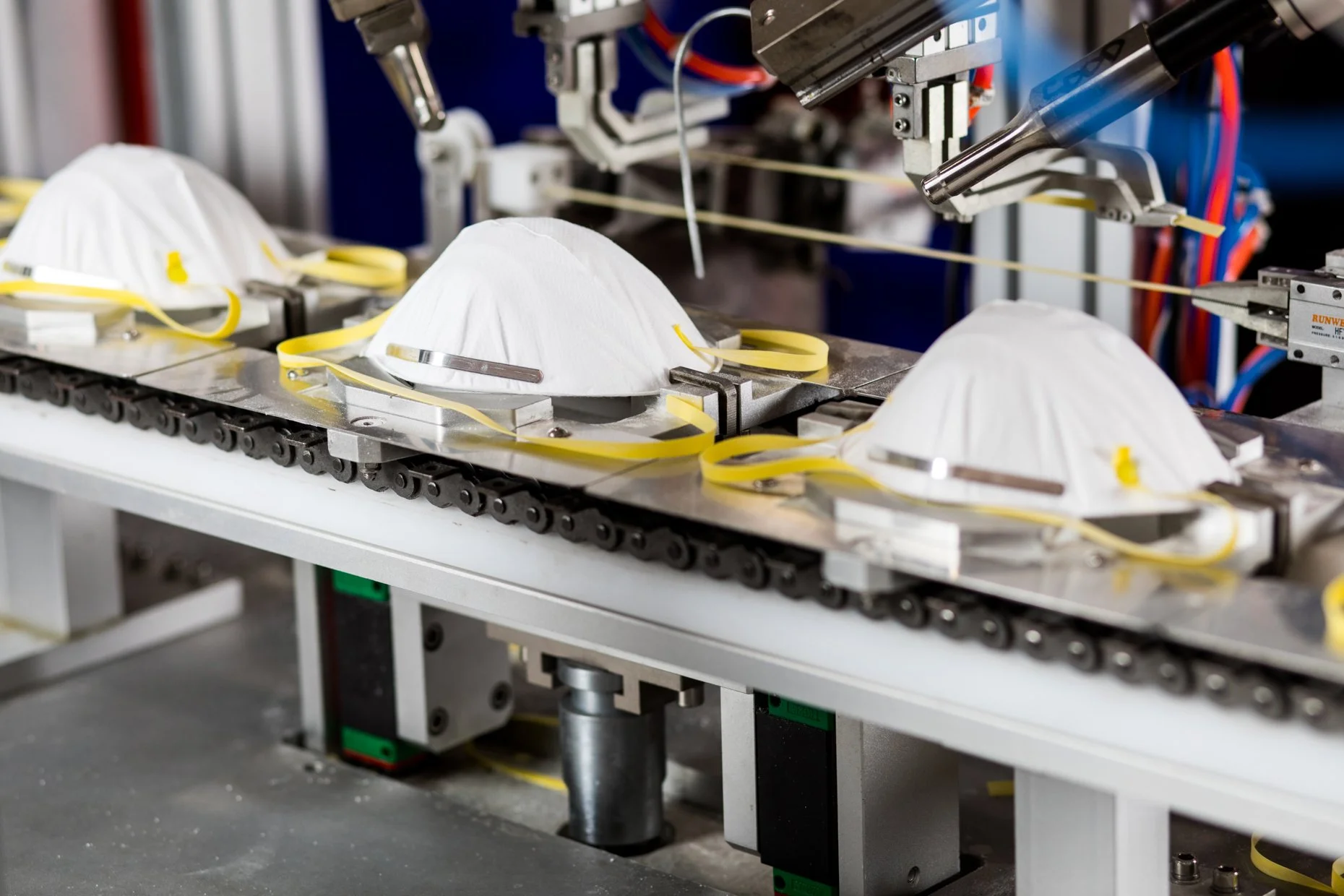 A manufacturing process involving robotic machinery assembling face masks on a conveyor belt, with several white masks lined up and connected by yellow straps.