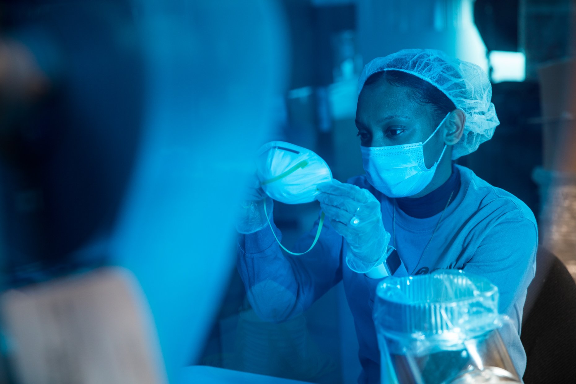 A woman wearing a hair cap, face mask, and gloves working in a laboratory, handling a piece of equipment or sample.