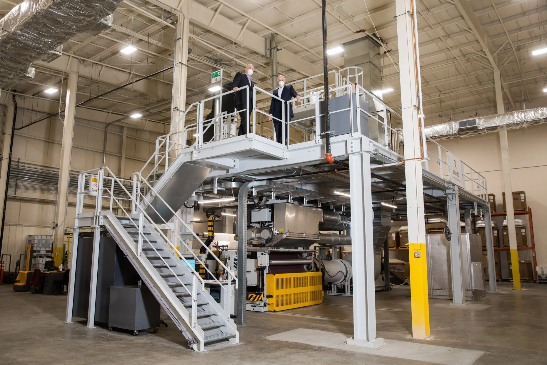 Two men in suits and face masks standing on an elevated platform inside an industrial warehouse, observing machinery and equipment below.