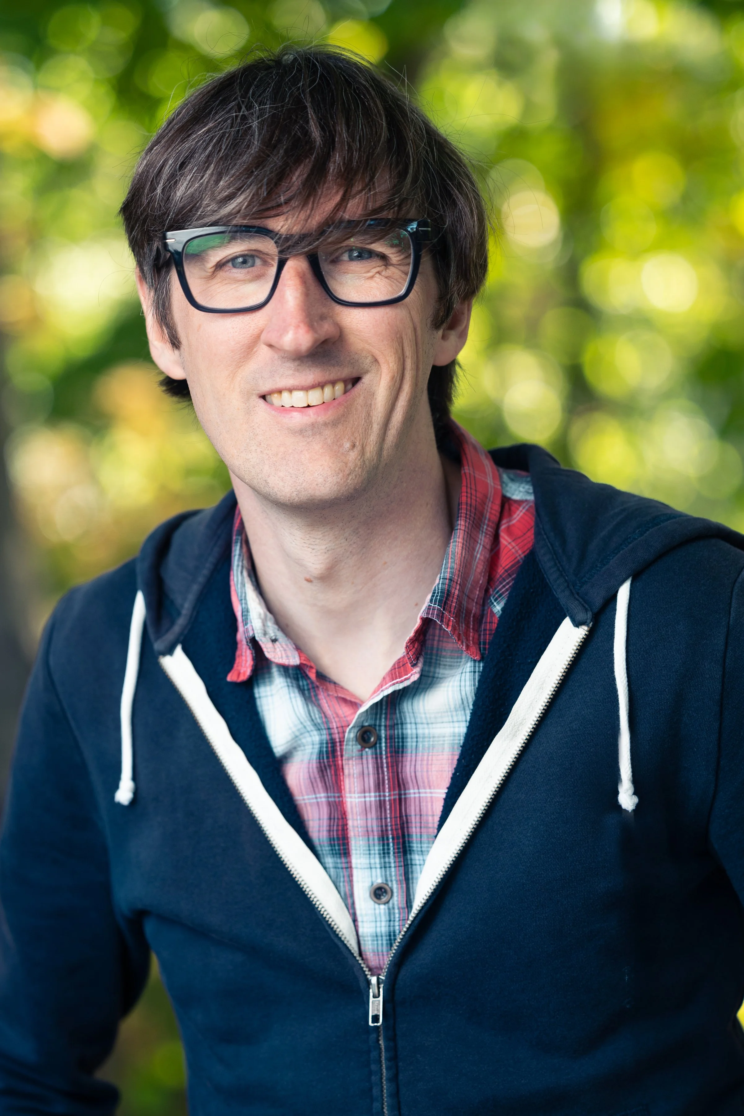 A man with dark brown hair, light blue eyes, and glasses smiling outdoors with green trees in the background. He's wearing a navy hoodie, a red plaid shirt, and a light blue shirt underneath.