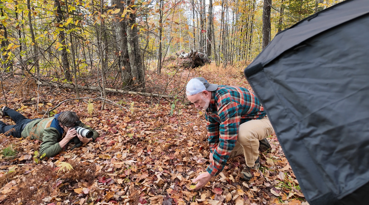 Two men are outdoors in a forest covered with fallen leaves during autumn. One man is lying on the ground, taking photos with a professional camera, while the other man is crouching, examining or collecting something from the leaves. A large black object, possibly part of a photography setup, is visible on the right.