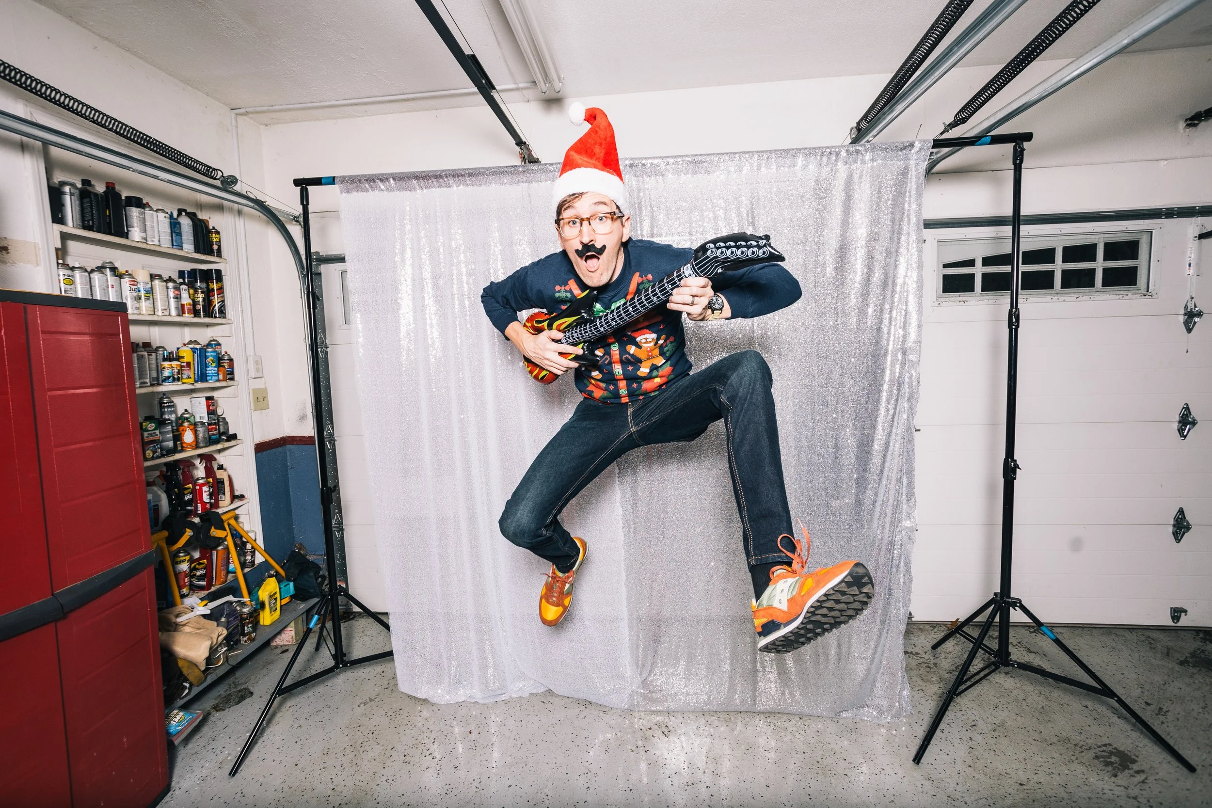 A man wearing a Santa hat, glasses, and a festive sweater is mid-air with an inflatable guitar, in a garage setting with scaffolding, tools, and a sparkle backdrop.
