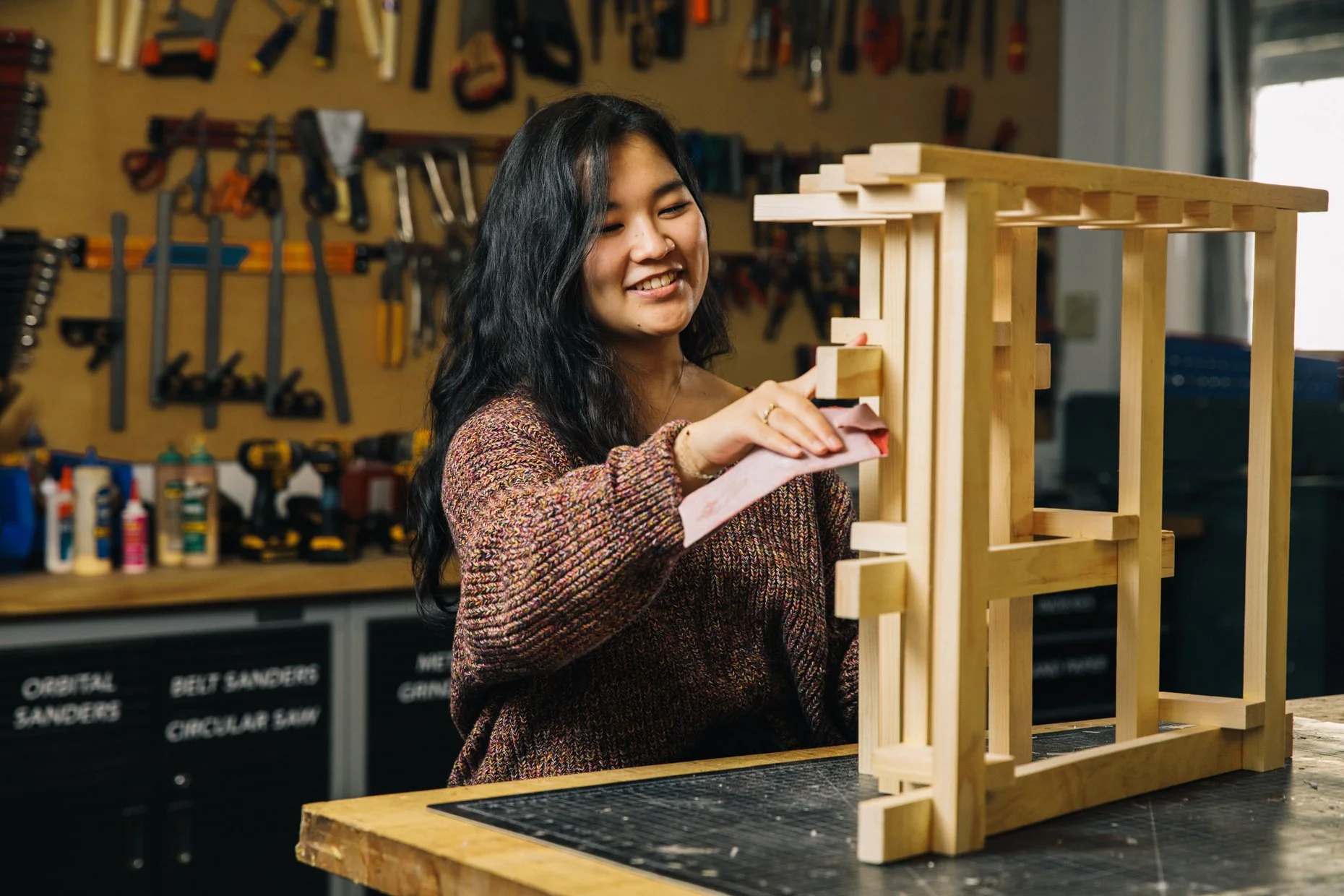 A young woman with long dark hair and a nose ring is sanding a wooden structure in a workshop. She is wearing a multicolored sweater and smiling. The workshop has various tools hanging on the wall and bottles of glue and other supplies on the workben