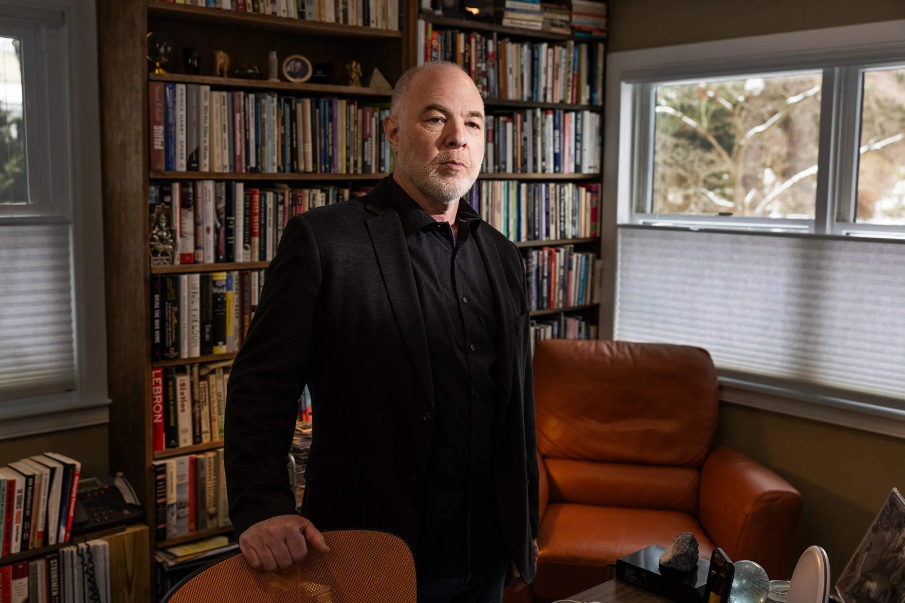 A middle-aged man with a beard in a black jacket standing in a room with bookshelves filled with books and a window showing a snowy outdoor scene.
