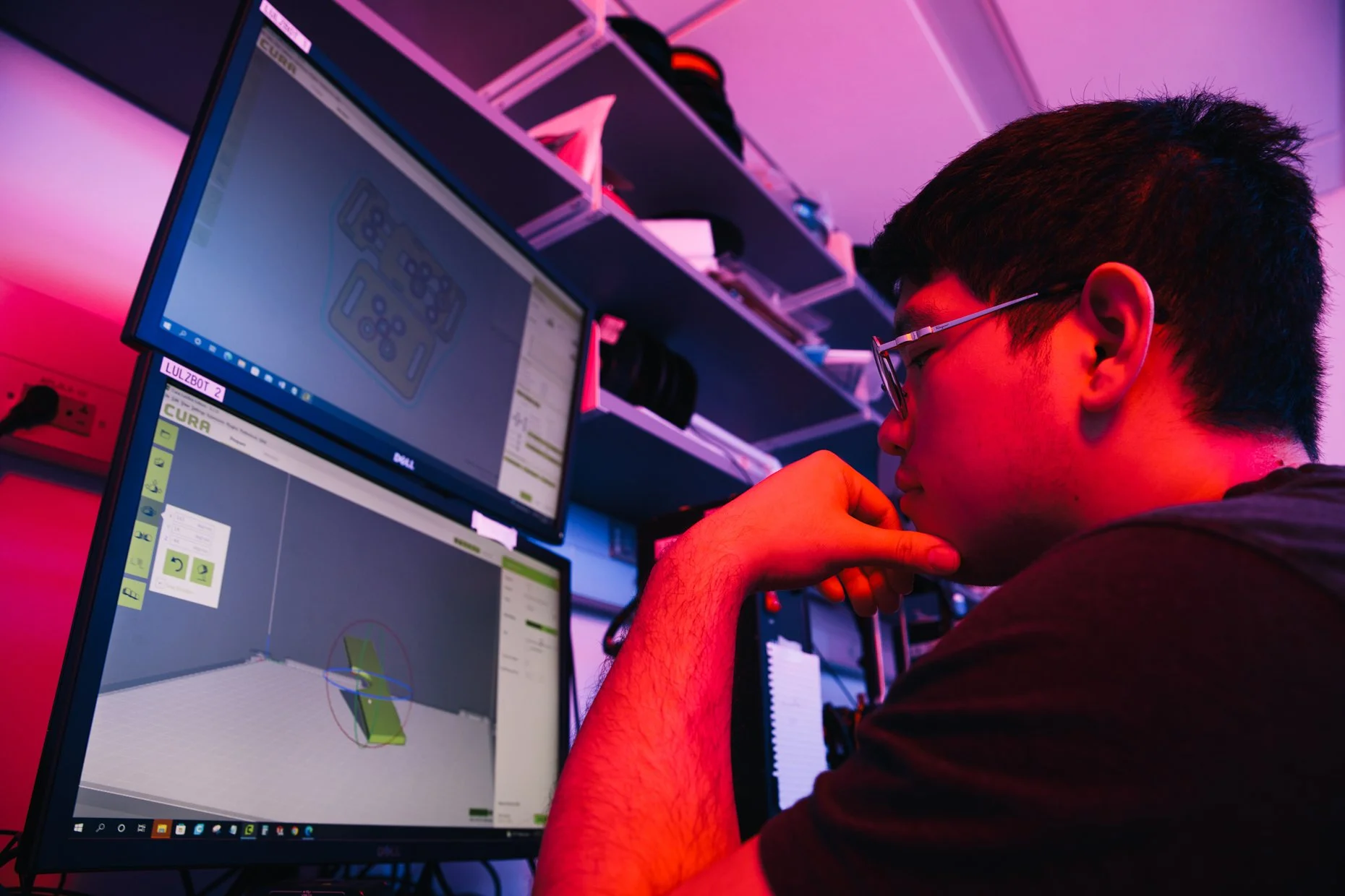 A person with glasses, sitting at a desk with multiple computer monitors displaying software interfaces, in a room with purple and pink lighting, appearing focused or deep in thought.