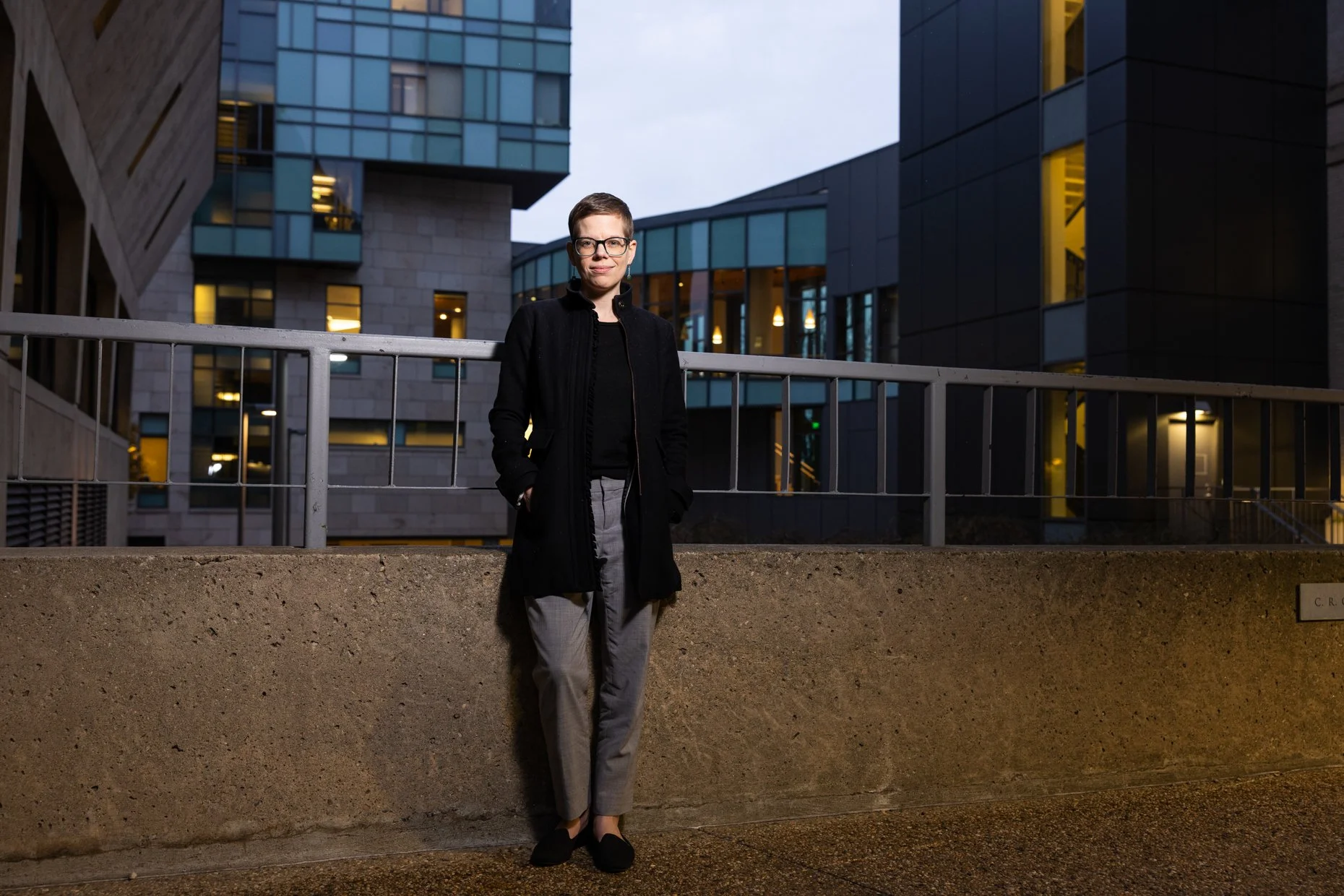 A woman with short brown hair, glasses, and earrings, wearing a black jacket, black top, gray pants, and black shoes, stands outdoors at dusk in front of a modern building with glass and concrete architecture, illuminated from within.