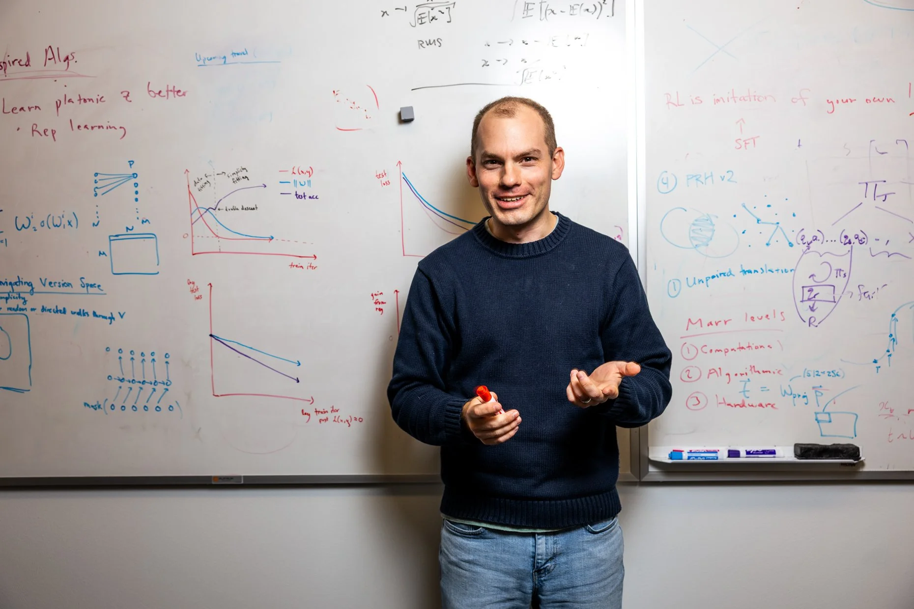 A man standing in front of a whiteboard filled with colorful handwritten notes and diagrams, smiling and holding a red marker.