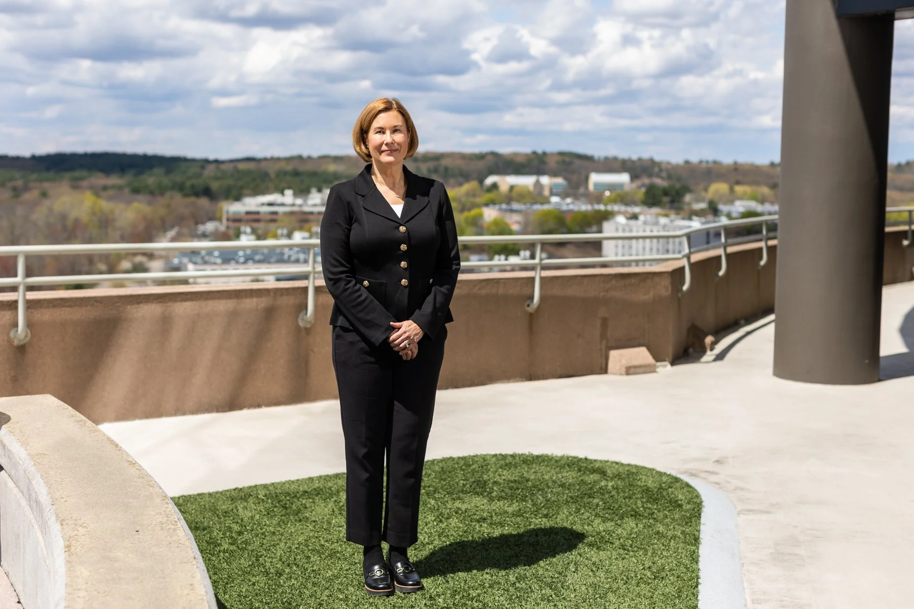 A woman in a black suit standing on a rooftop terrace with a cityscape and mountains in the background.
