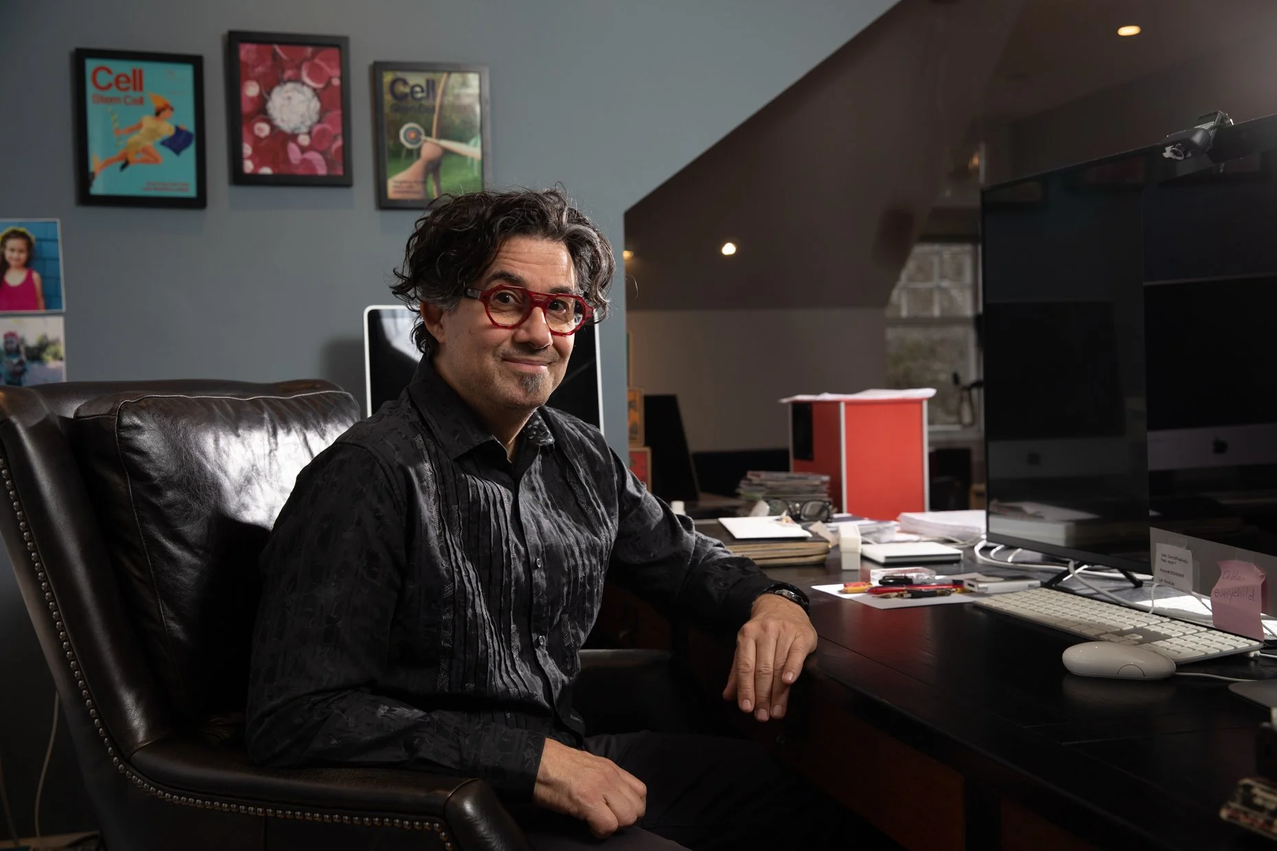 A man with curly dark hair, red glasses, and a black patterned shirt sitting at a cluttered desk in a home office, smiling at the camera. The background features framed artwork on the wall, a computer monitor, and a window.