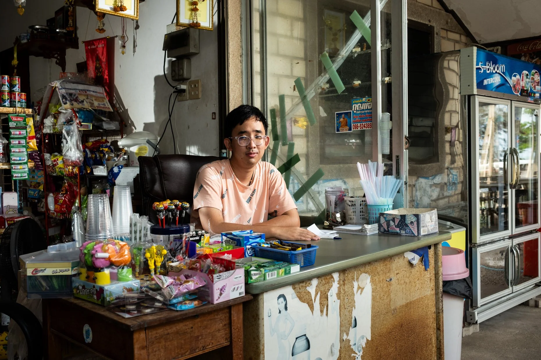 A young man with glasses sitting behind a counter in a small store, surrounded by snacks, candies, and plastic cups, with a refrigerator and shelves in the background.