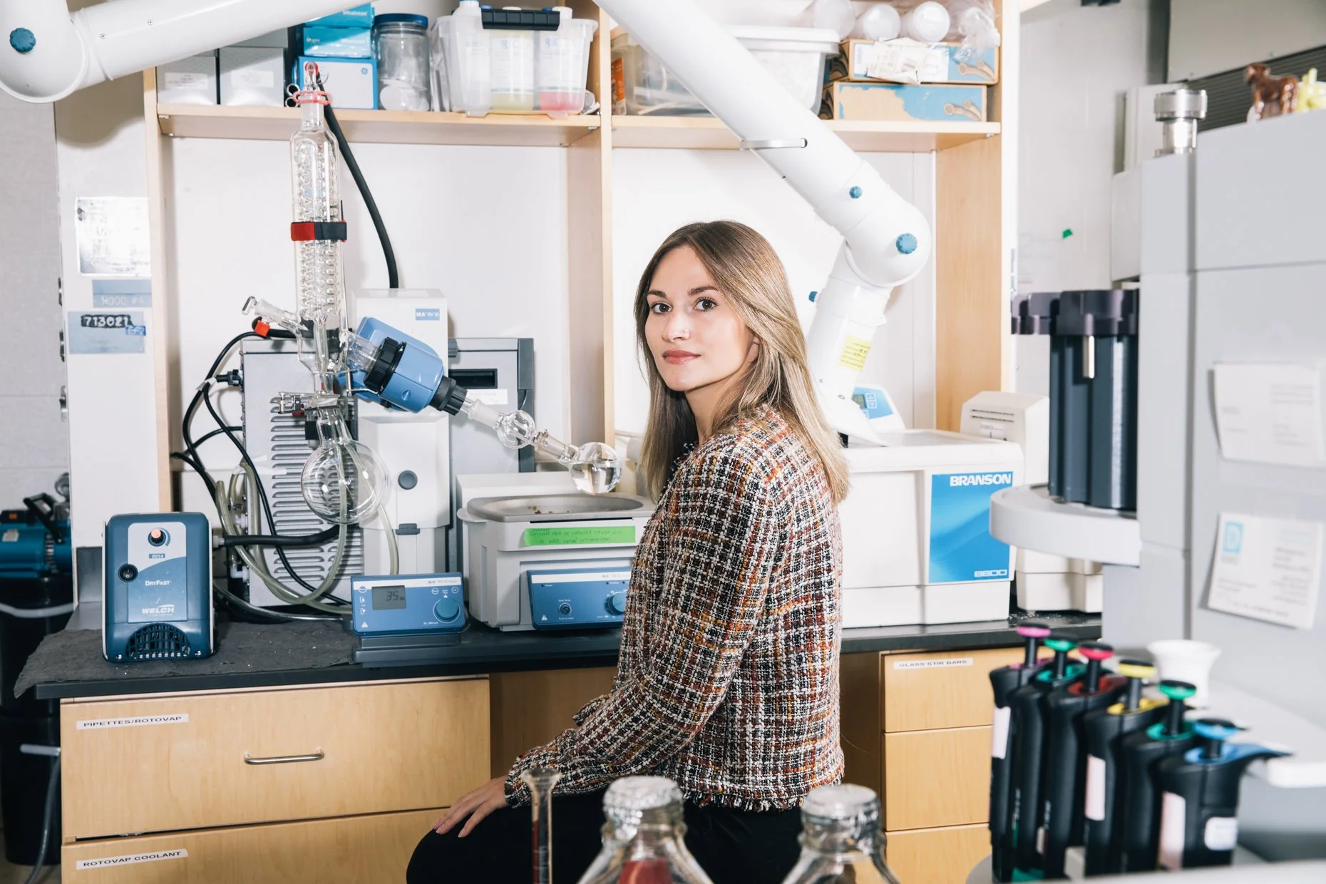 A woman in a lab coat sitting in a chemistry laboratory surrounded by scientific equipment, glassware, and lab instruments.