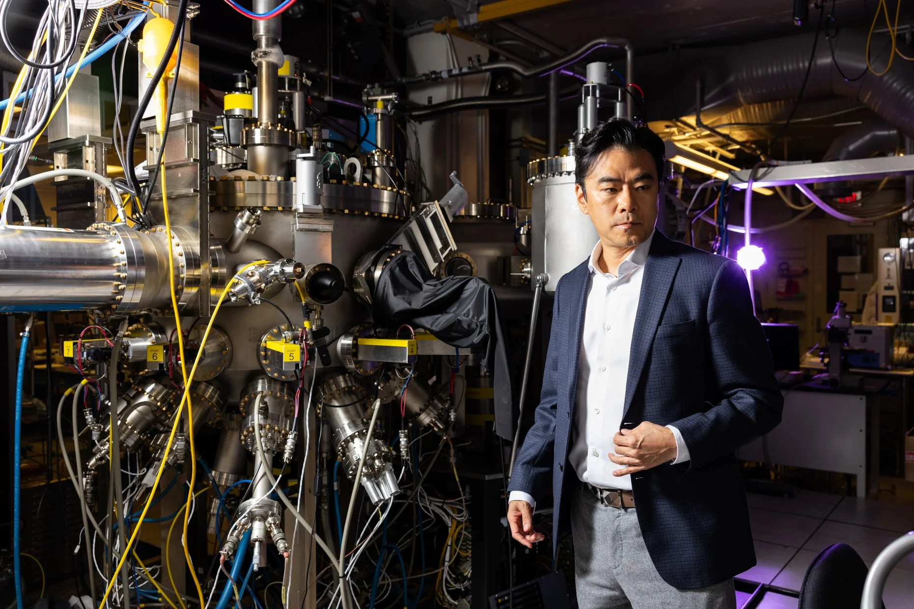 A man in a business suit and white shirt stands in a laboratory or industrial setting with complex machinery and numerous wires and tubes, appearing to be inspecting or observing the equipment.