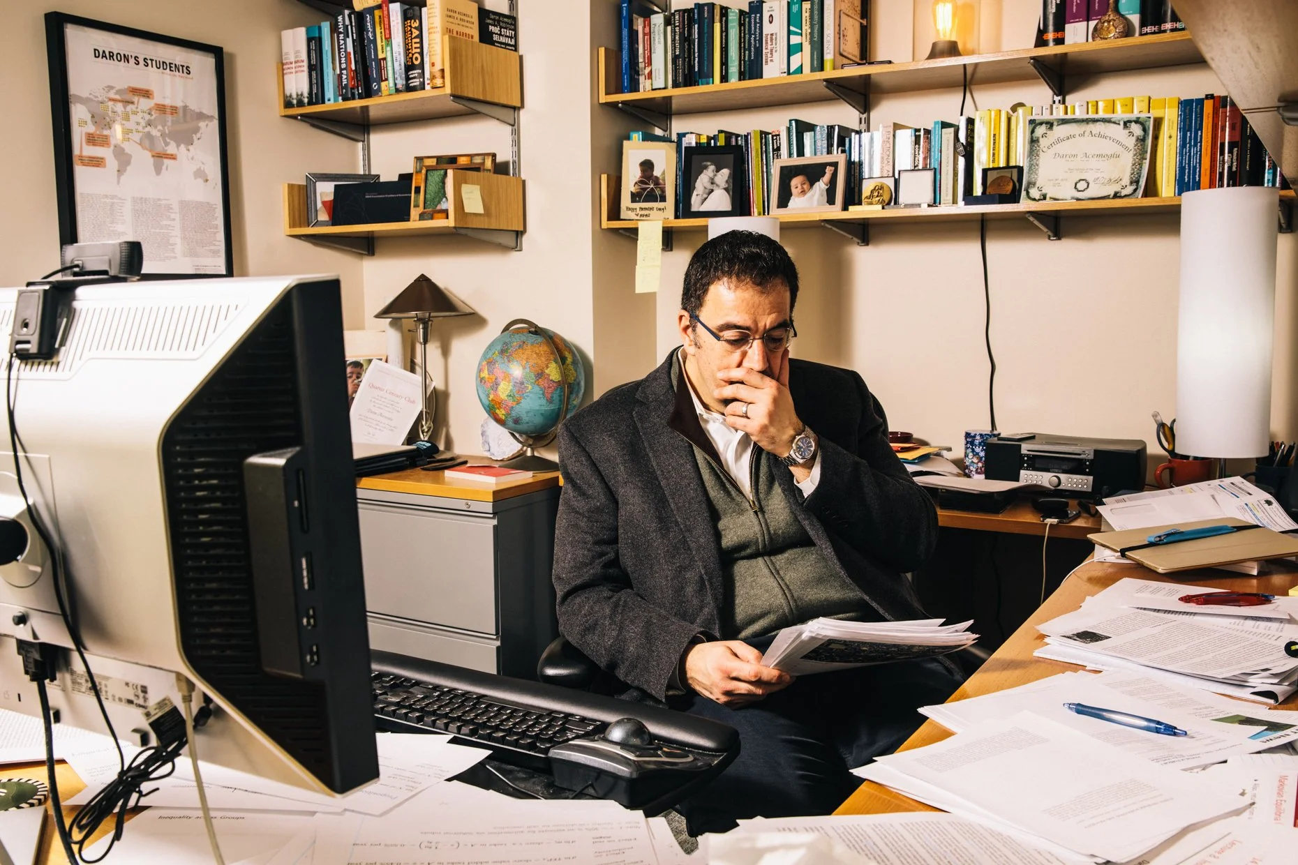 A man sitting at a cluttered desk in a home office, reading a document. The desk has papers, a computer, and a pen. Behind him are shelves filled with books, framed photos, and awards. A small globe and a desk lamp are visible nearby.
