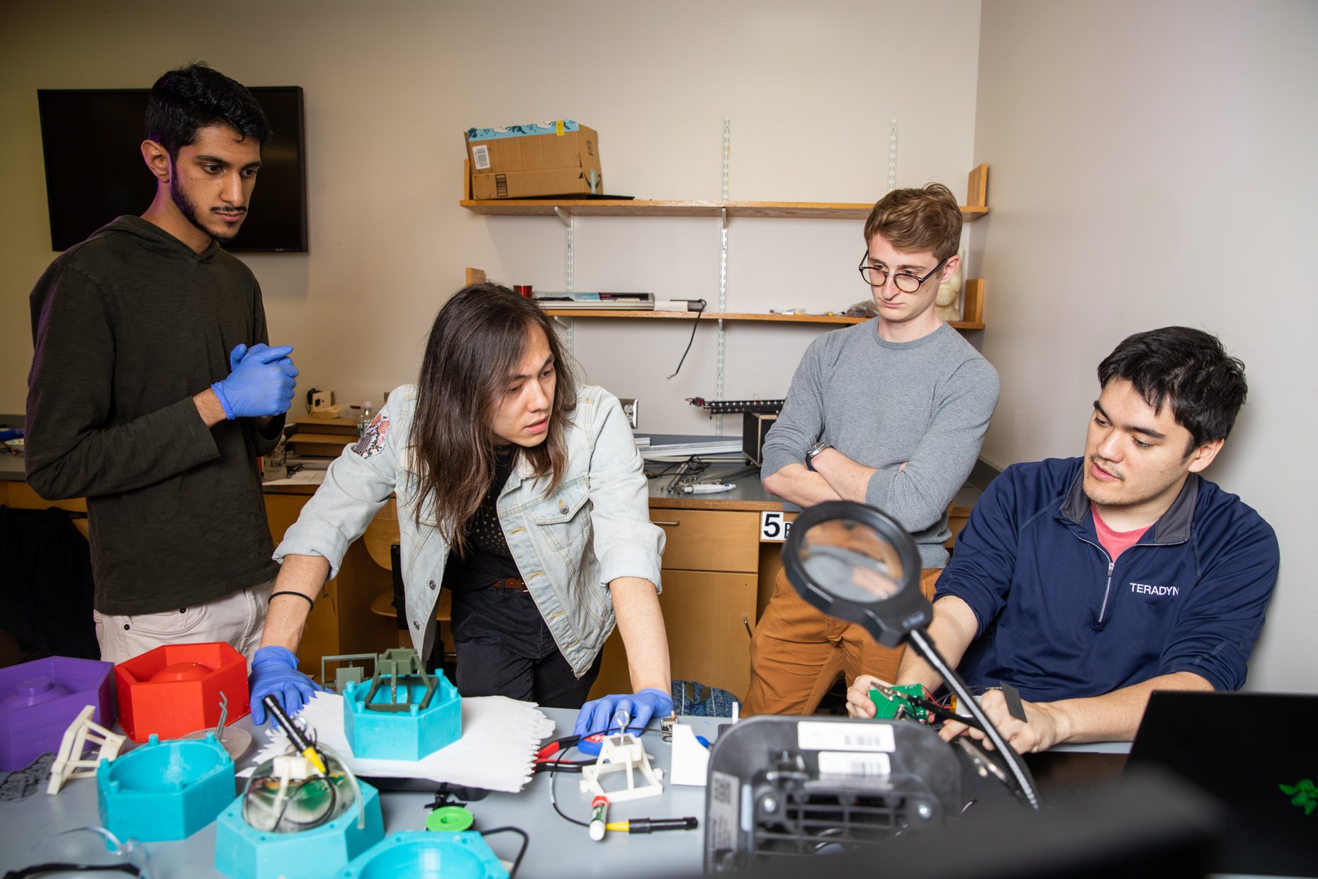 Five people working on a project in a laboratory or workshop setting, with various electronic components and tools on the table.