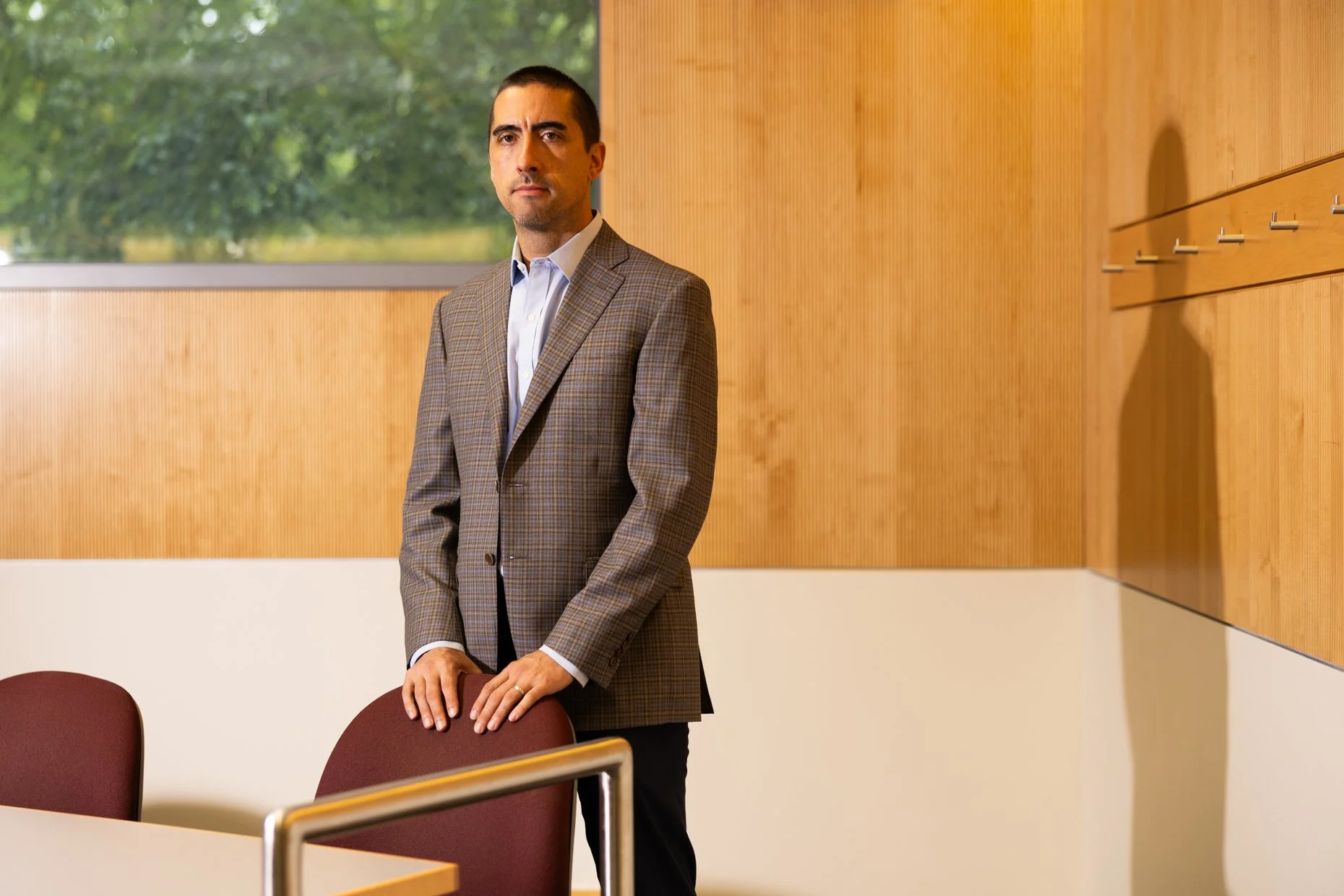 A man in a plaid suit and white shirt stands in a conference room, holding the back of a maroon chair. The room has a window with green trees visible outside, wood paneling on the wall, and a table with several chairs.