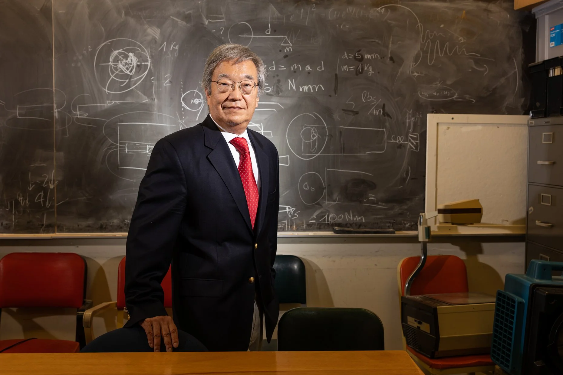 A distinguished older man in a dark blazer, white shirt, red tie, and glasses standing in a classroom in front of a chalkboard filled with scientific and mathematical drawings and equations. He is standing next to chairs and a desk, with a chest of d
