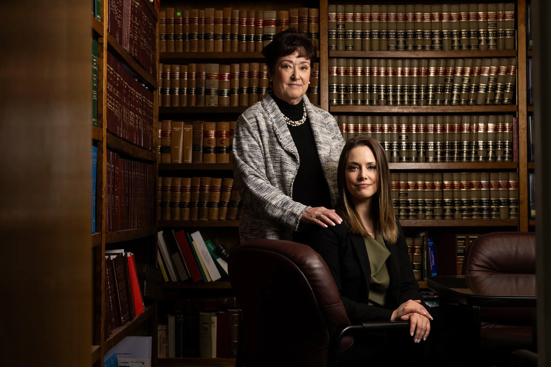Two women in a law library with wooden shelves filled with law books. One woman stands behind the other, who is seated, both looking at the camera.