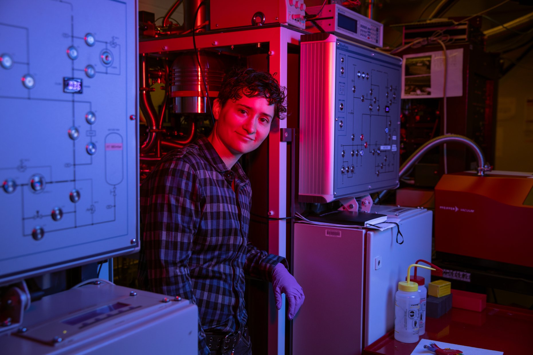 A young man in a checkered shirt and white gloves working in a laboratory with scientific equipment, illuminated by red and purple lighting, surrounded by scientific instruments and laboratory bottles.