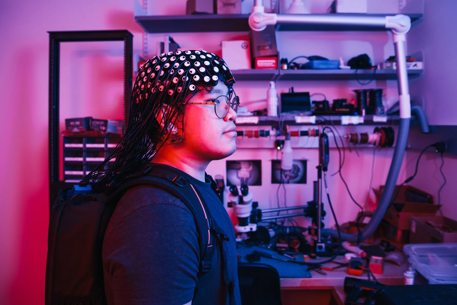 Man wearing an EEG cap with multiple electrodes connected to wires, in a lab with technical equipment and electronic tools on shelves, illuminated with purple and pink lighting.