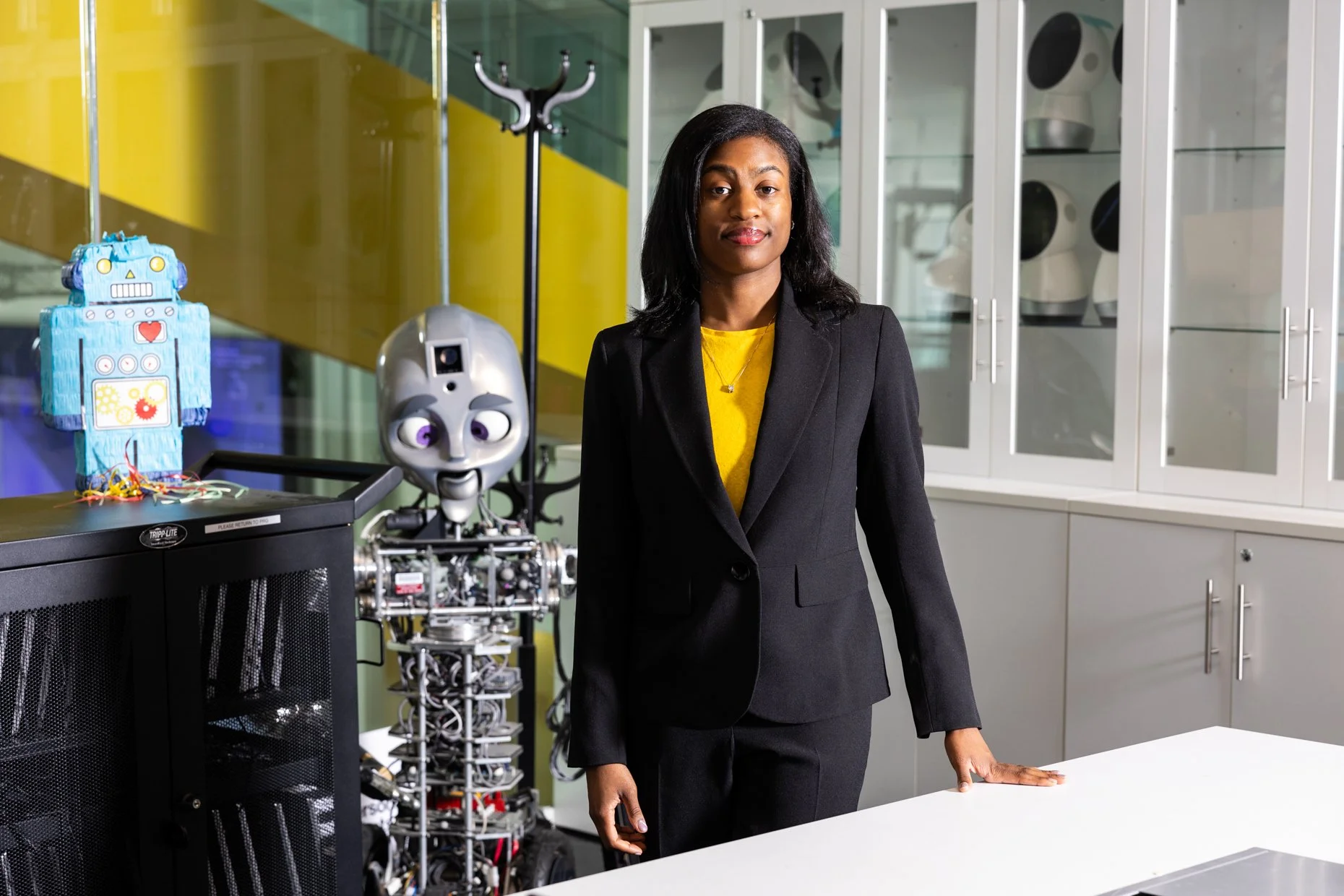 A woman in a black blazer and yellow shirt standing in a high-tech laboratory with robotic prototypes and testing equipment visible behind her.