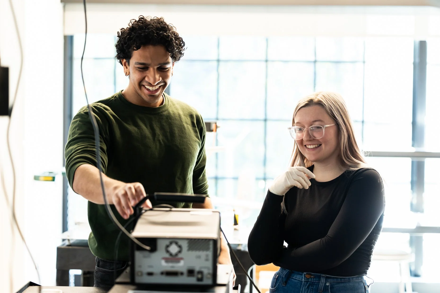 Two people, a man and a woman, working together on a technical project in a laboratory or workshop. The man is smiling and pointing at a device, while the woman, wearing glasses and gloves, is attentively looking at the device.