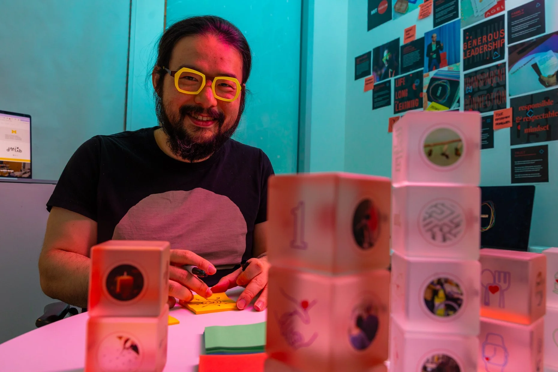 A man with long black hair, glasses, and a beard, smiling at the camera, sitting at a table with colorful blocks and paper, in a room with blue walls and posters in the background.