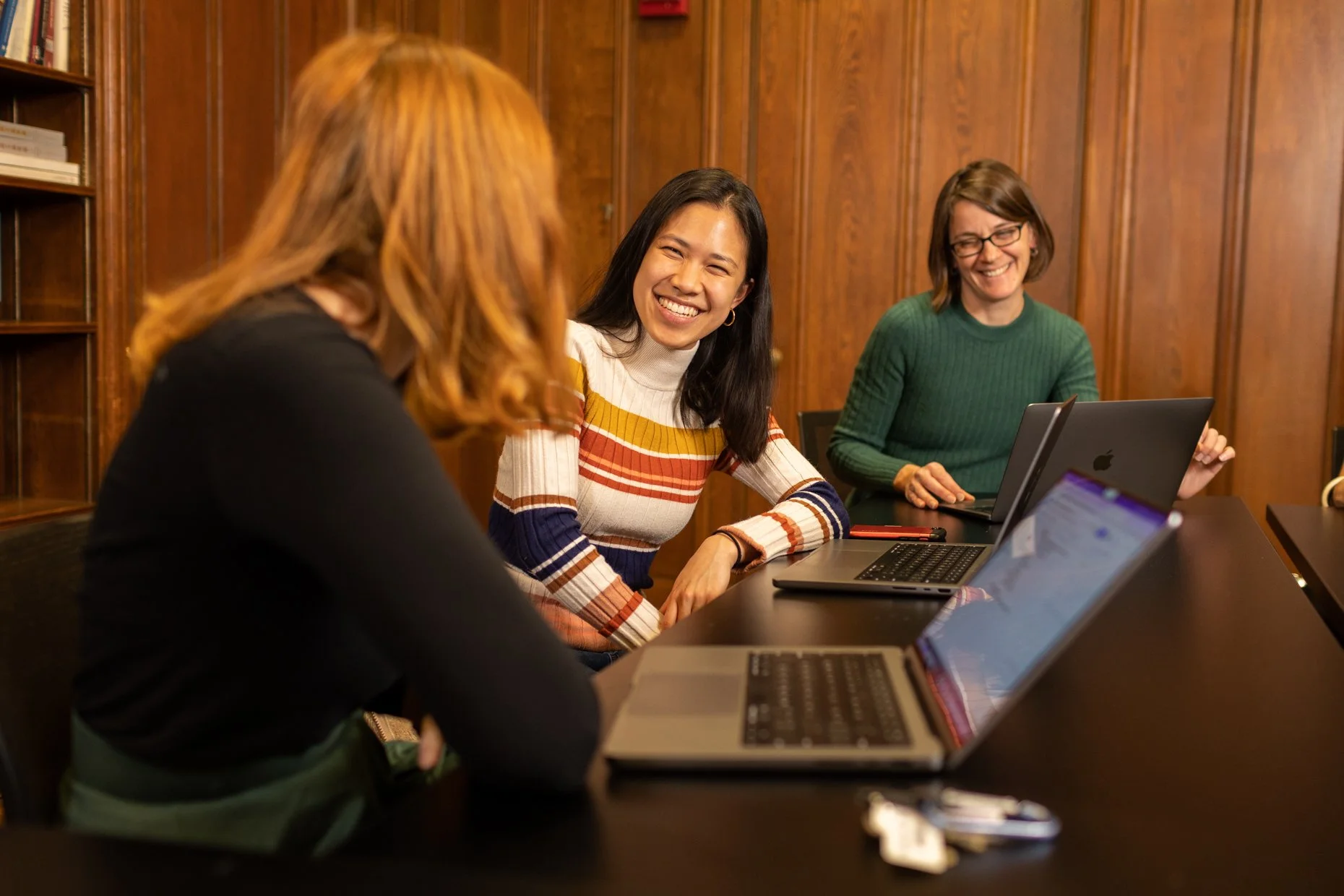 Three women sitting at a conference table with laptops, engaged in conversation in a wood-paneled room. One woman is smiling broadly, wearing a striped sweater, while another woman with glasses and a green sweater is smiling.