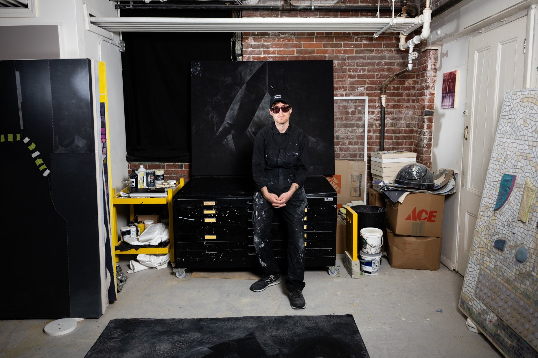 A man wearing sunglasses, a black cap, and black overalls stands in an art studio or workshop, surrounded by paint supplies, cardboard boxes, and artwork against a brick wall. He is sitting on a black cabinet covered in paint splatters.