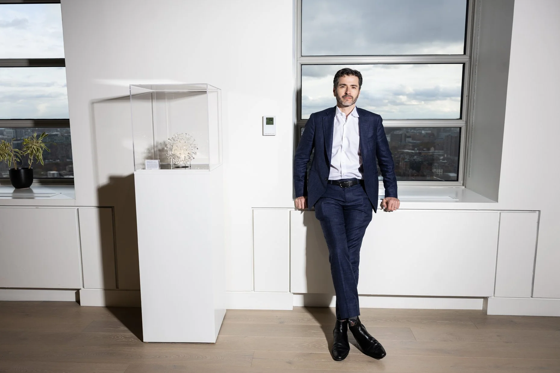 A man in a dark blue suit and black boots stands leaning against a windowsill in a modern, white room with large windows. The cityscape and cloudy sky are visible outside.