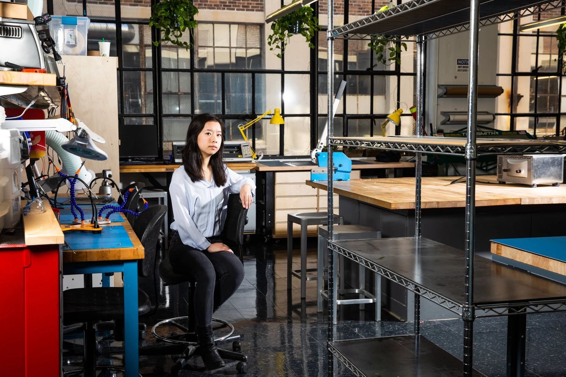 A woman sitting in an industrial-style workshop or laboratory, surrounded by workbenches, shelves, tools, and equipment, with large windows and exposed brick walls in the background.