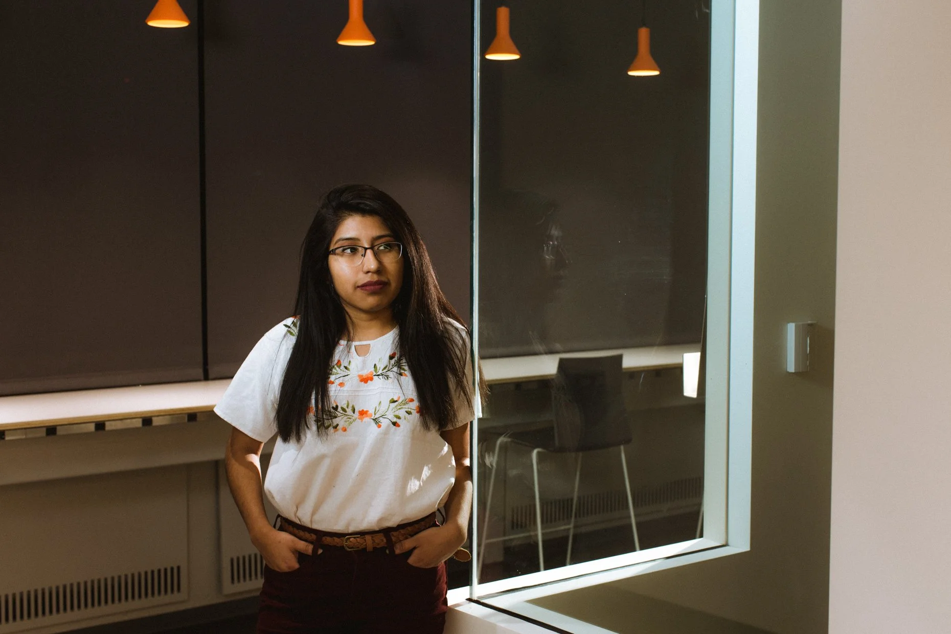 A woman with long dark hair, glasses, and a white embroidered blouse standing indoors near a glass window, with a dark wall and orange pendant lights above.