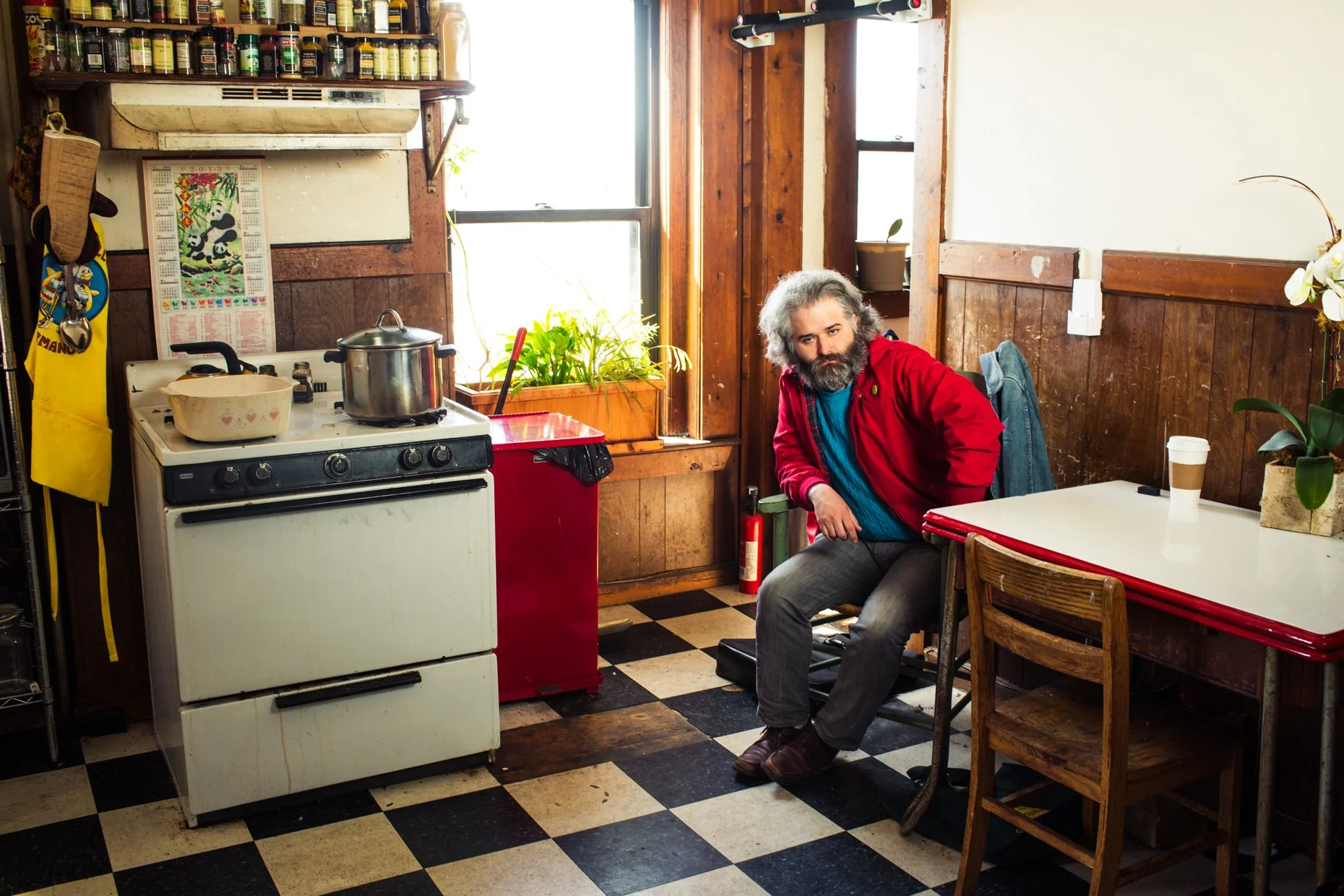 A man with gray hair and beard sitting at a table in a rustic kitchen with wood-paneled walls, black-and-white checkered floor, and a window. There are kitchen appliances, potted plants, and a coffee cup on the table.
