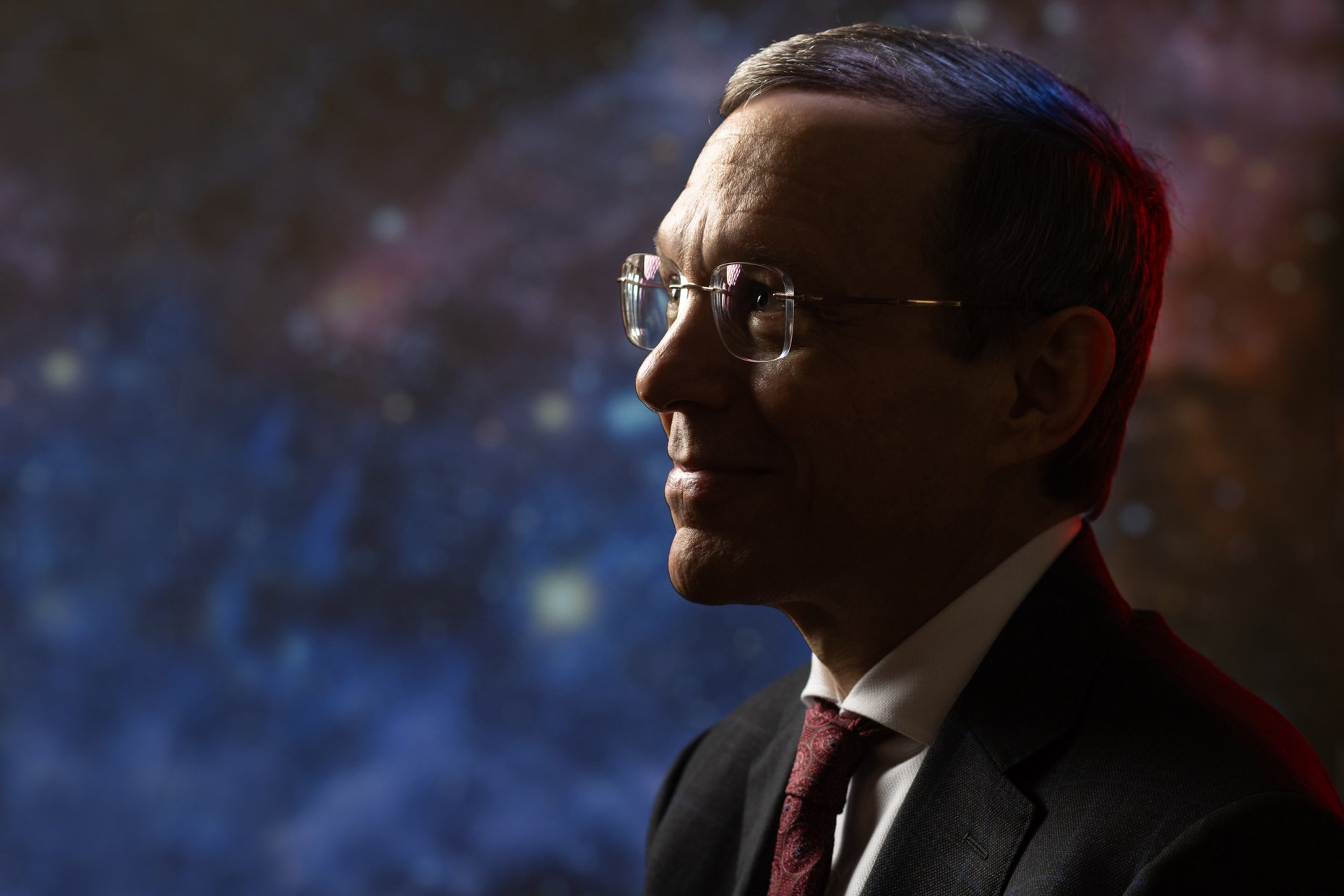 A profile of a man wearing glasses, a dark suit, white shirt, and red tie, with short dark hair, illuminated by colorful lighting against a blurred, colorful background.