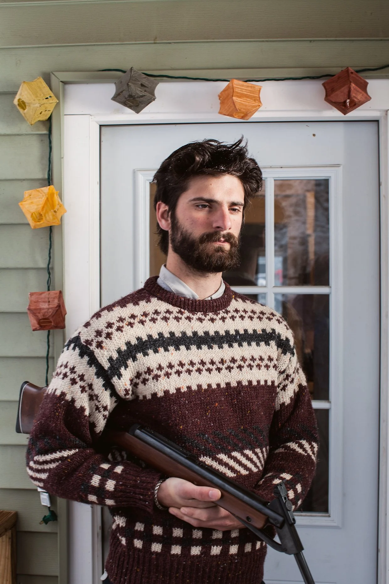 A man with dark hair and beard wearing a patterned sweater, holding a shotgun, standing outside near a glass door with string lights decorated with colorful paper lanterns.