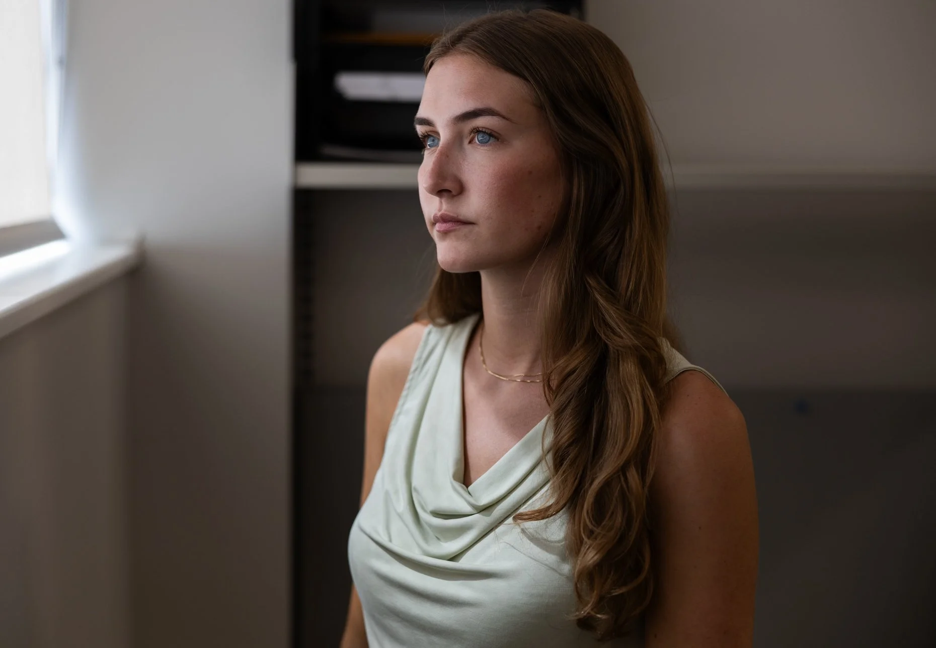 A woman with long wavy brown hair and blue eyes looking out a window, wearing a sleeveless cream-colored top and a gold necklace, with a neutral expression in a softly lit room.